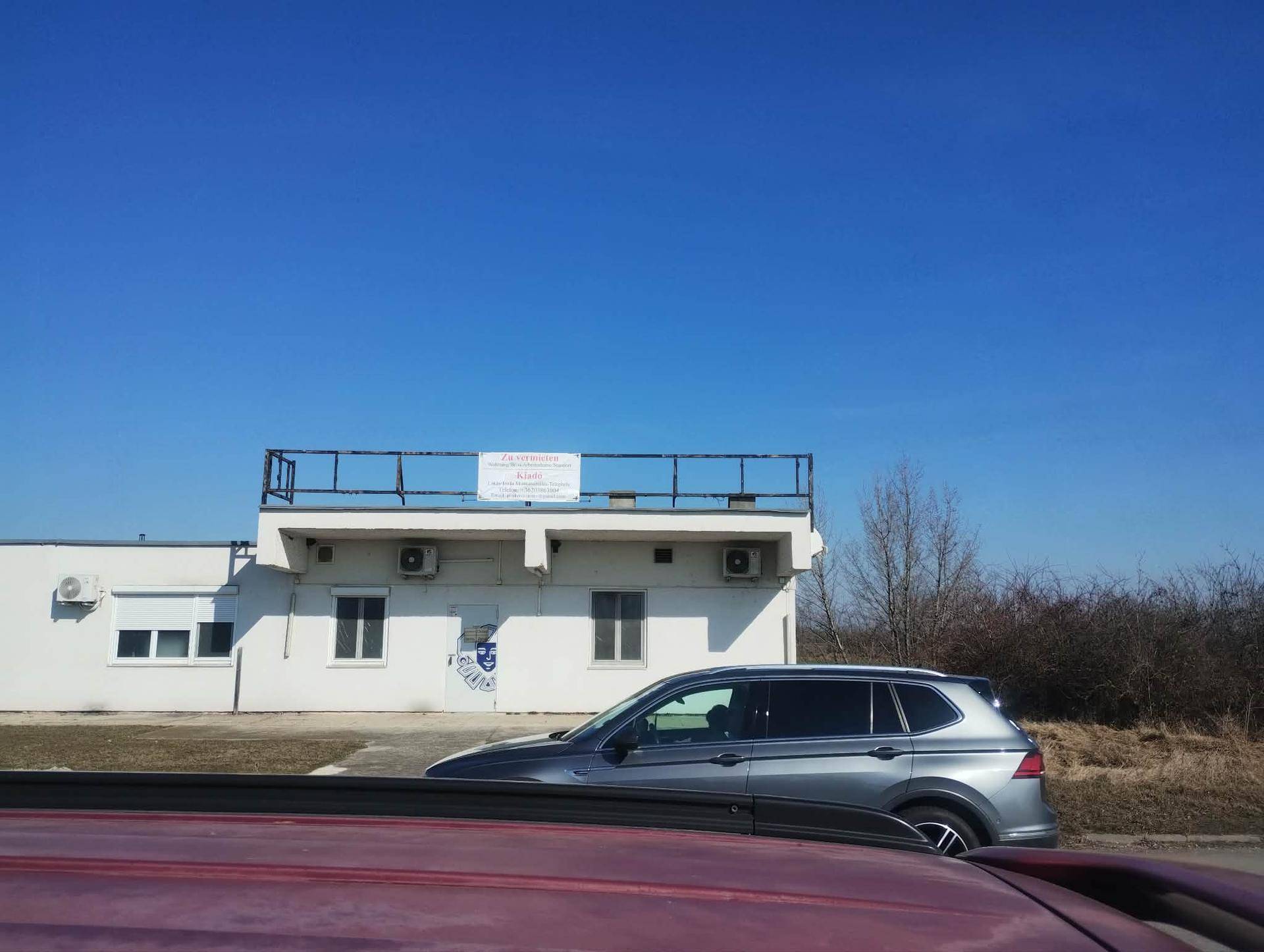 Außenansicht eines weißen Gebäudes mit Flachdach und Geländer, teilweise verdeckt von einem Auto.