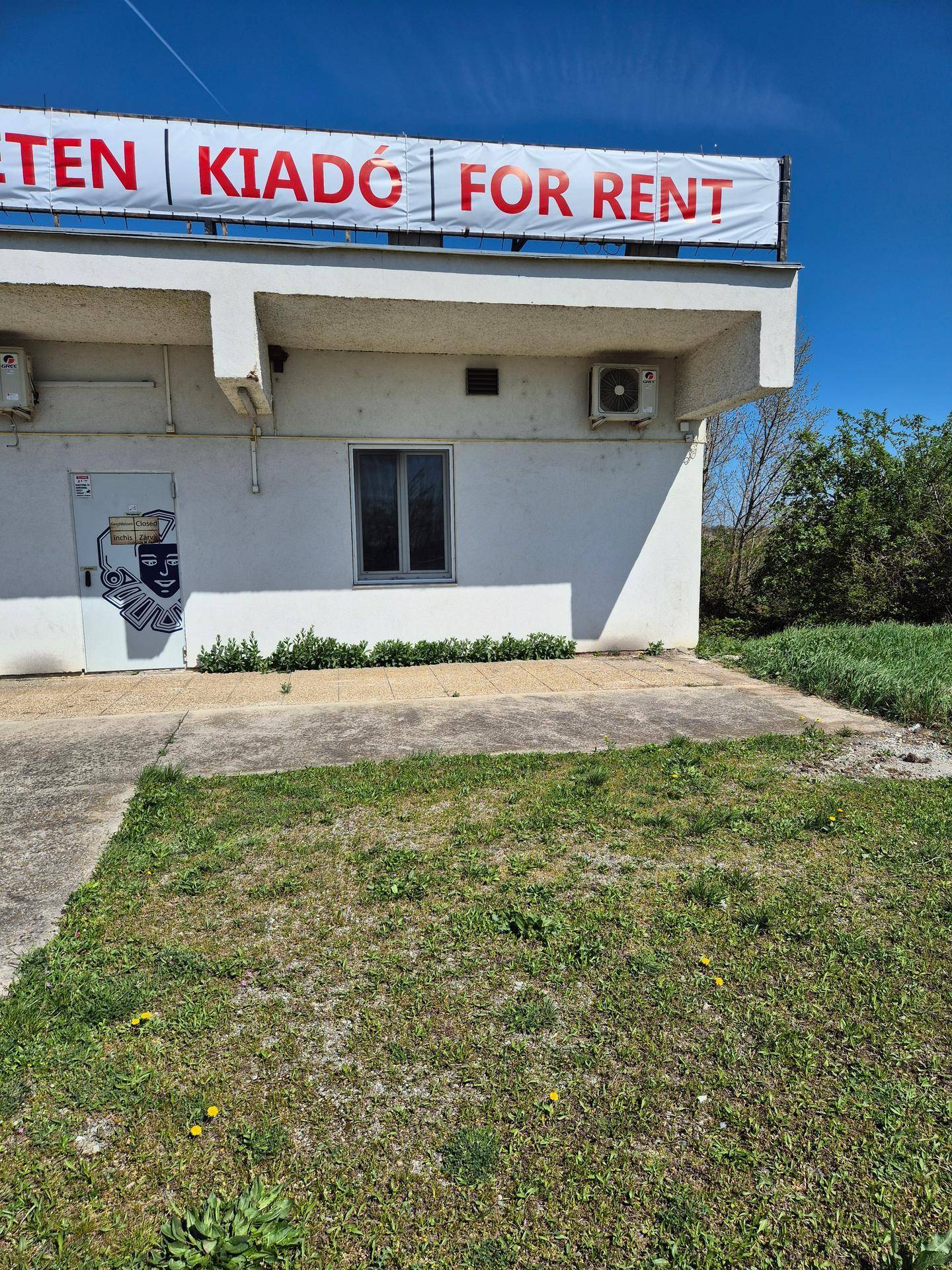 Nahaufnahme der weißen Gebäudefassade mit einem 'FOR RENT' Banner auf dem Dach und einer kleinen Grünfläche.