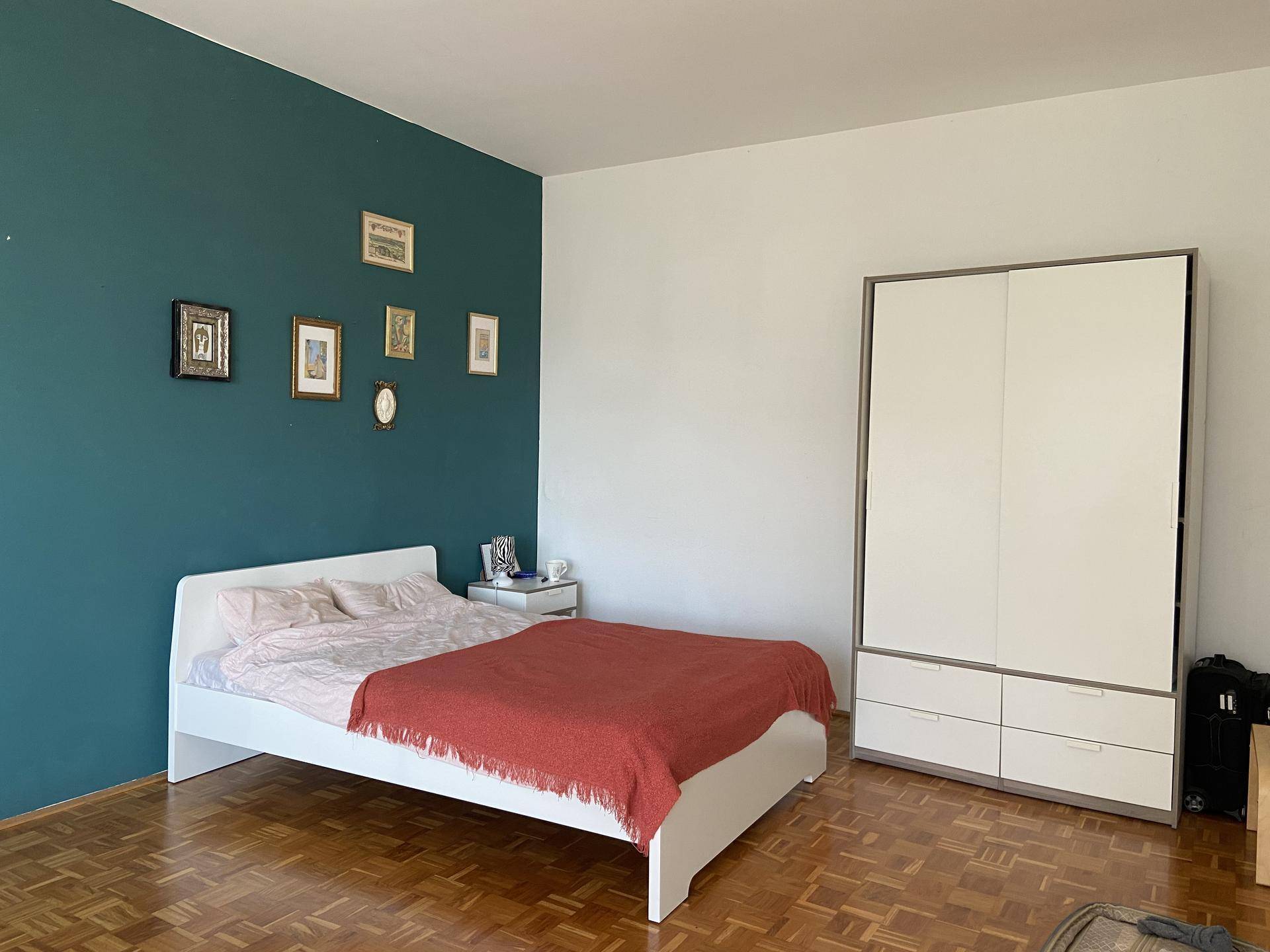 Schlafzimmer mit tealfarbener Wand, weißem Bett und modernem Kleiderschrank auf Parkettboden.