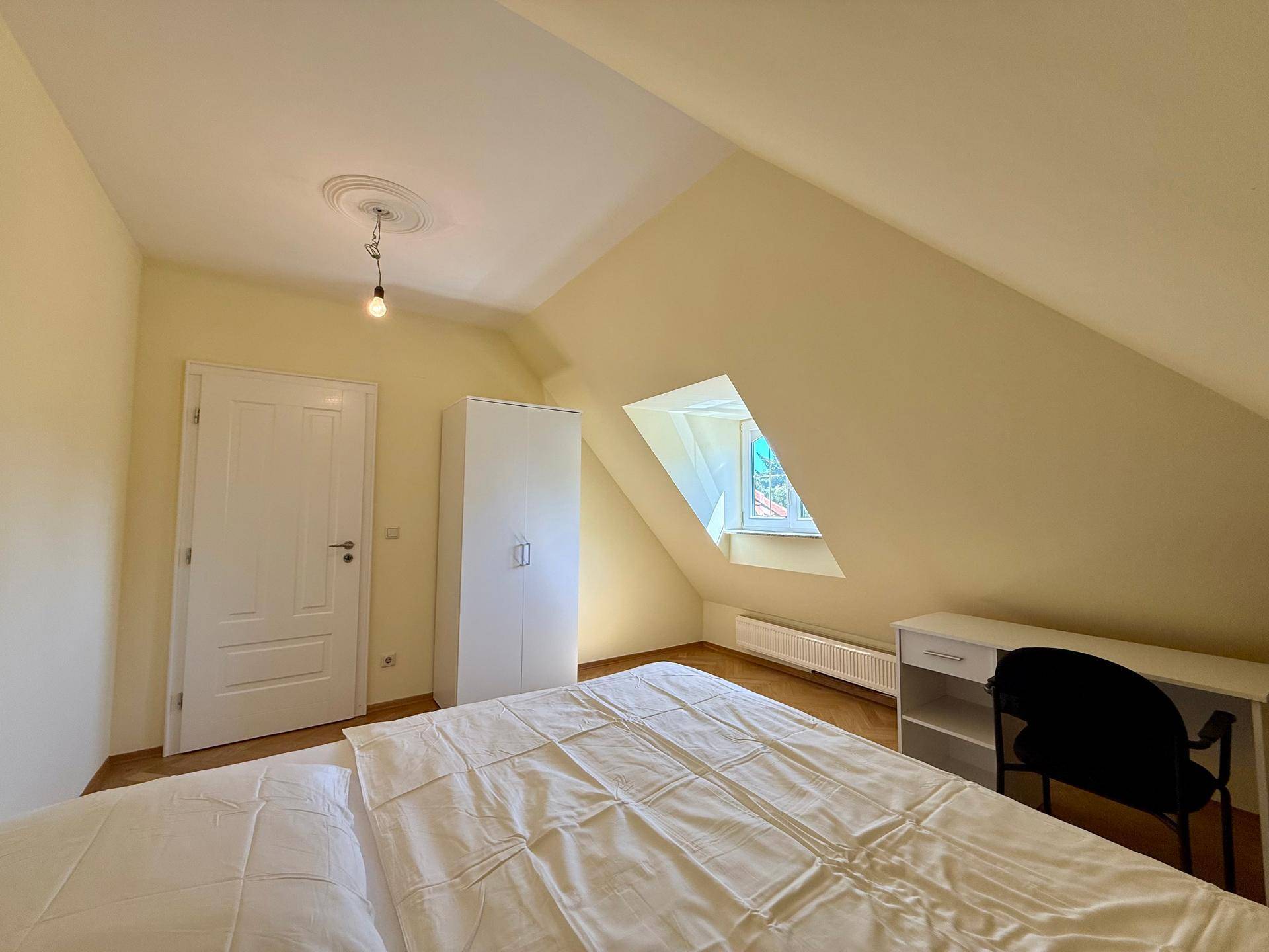 Dachgeschoss-Schlafzimmer mit Bett, Kleiderschrank und Schreibtisch, hell durch das Dachfenster.