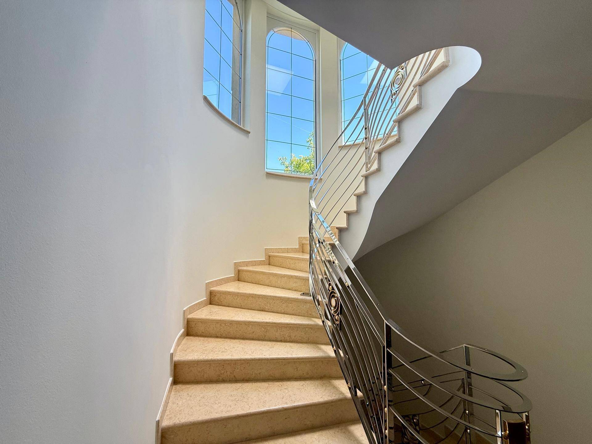 Elegante Wendeltreppe mit Steinstufen und Metallgeländer, beleuchtet durch große Bogenfenster.