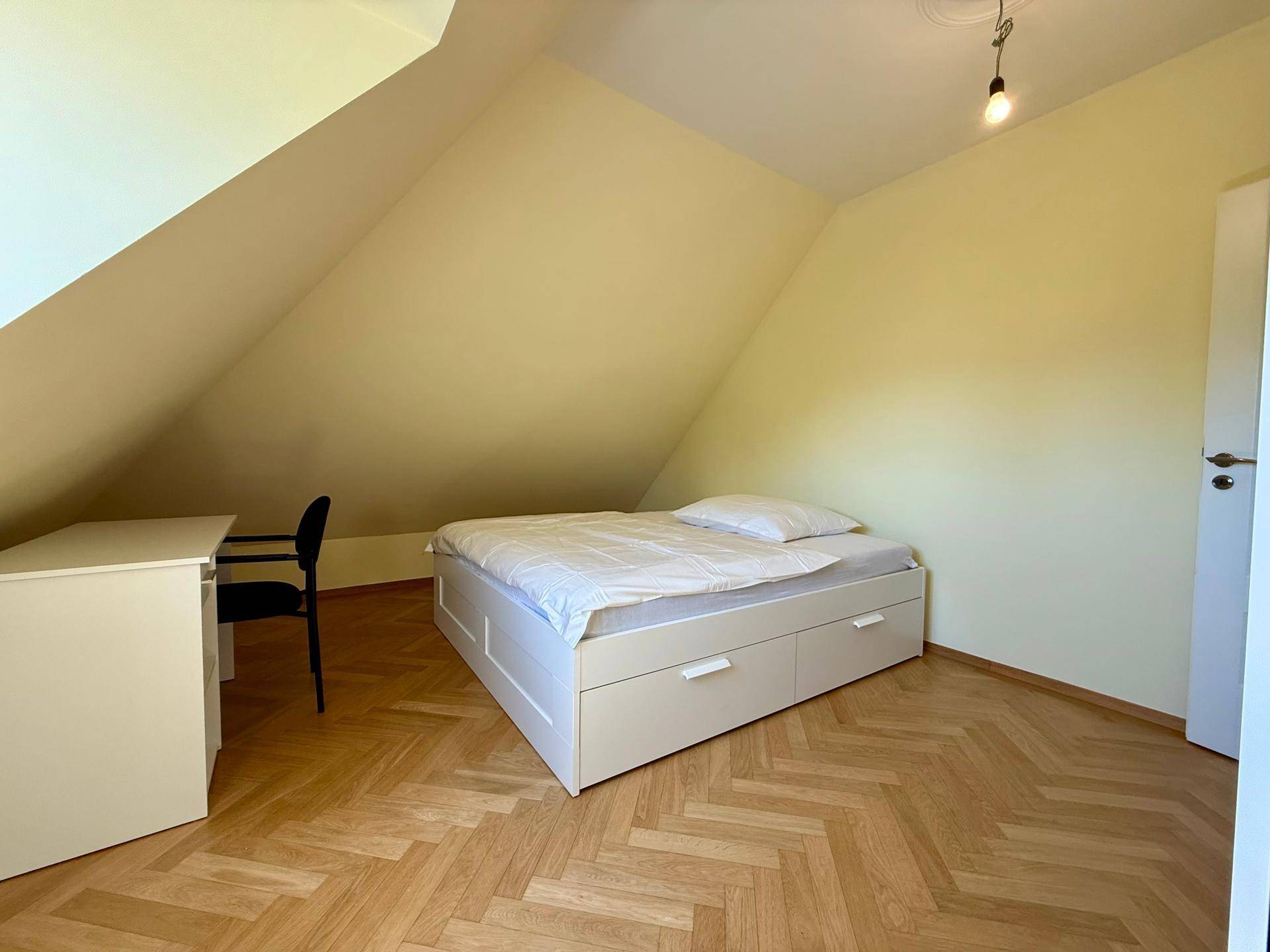 Schlafzimmer mit Dachschrägen, Parkettboden und einem einfachen Bett mit integriertem Stauraum.