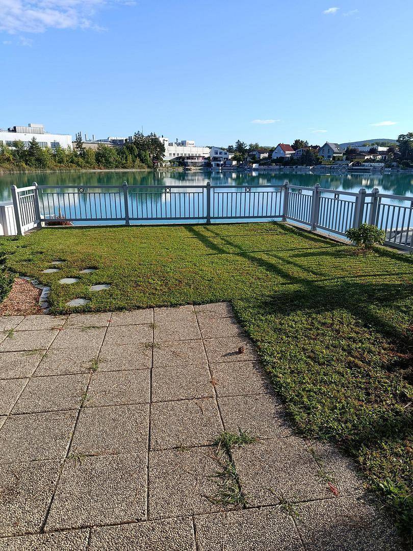 Rasenfläche mit gepflastertem Bereich und Zaun, der den Blick auf den See freigibt.