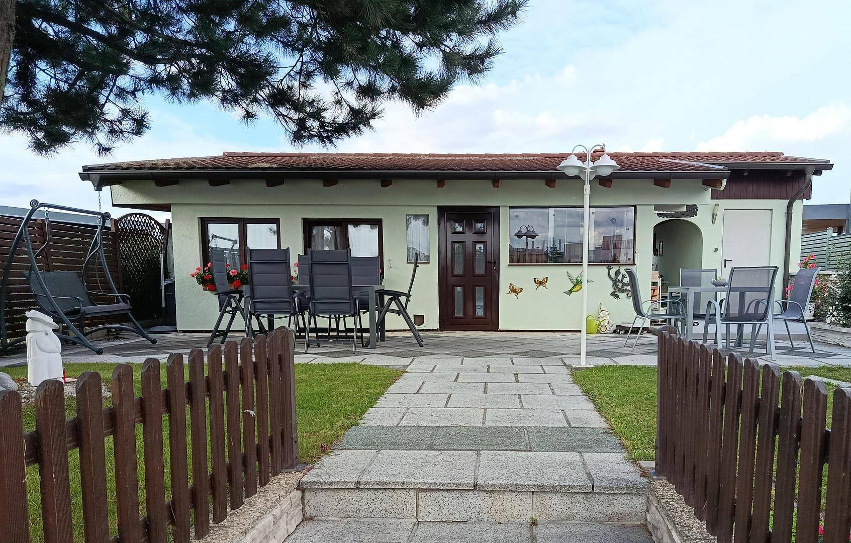 Kleines Haus mit Terrasse und Gartenbereich, umgeben von Grün und einem Holzzaun.