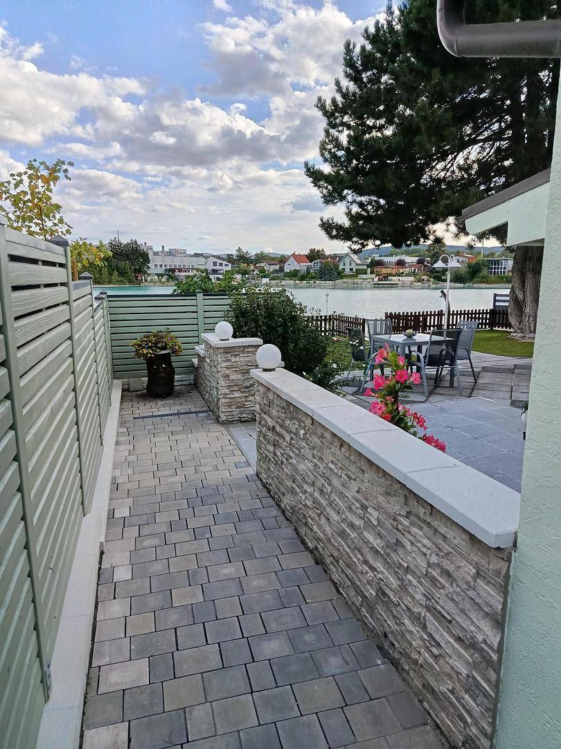 Gepflasterter Weg neben dem Haus führt zu einer Terrasse mit Seeblick und Blumen.