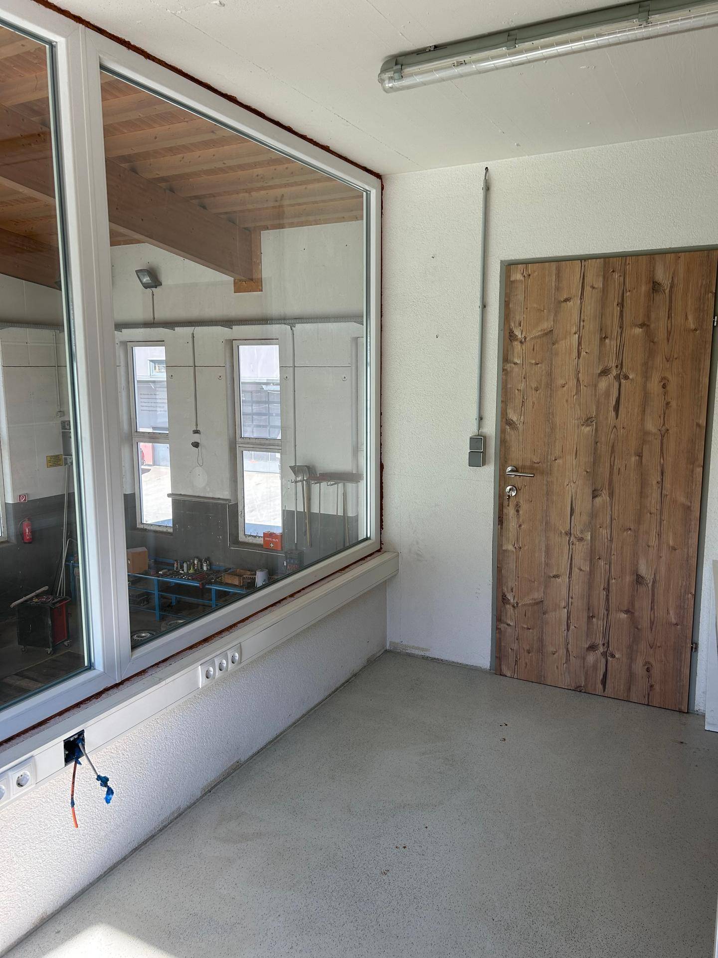 Büroraum im Obergeschoss mit großem Fenster zur Werkstatt und einer Holztür.