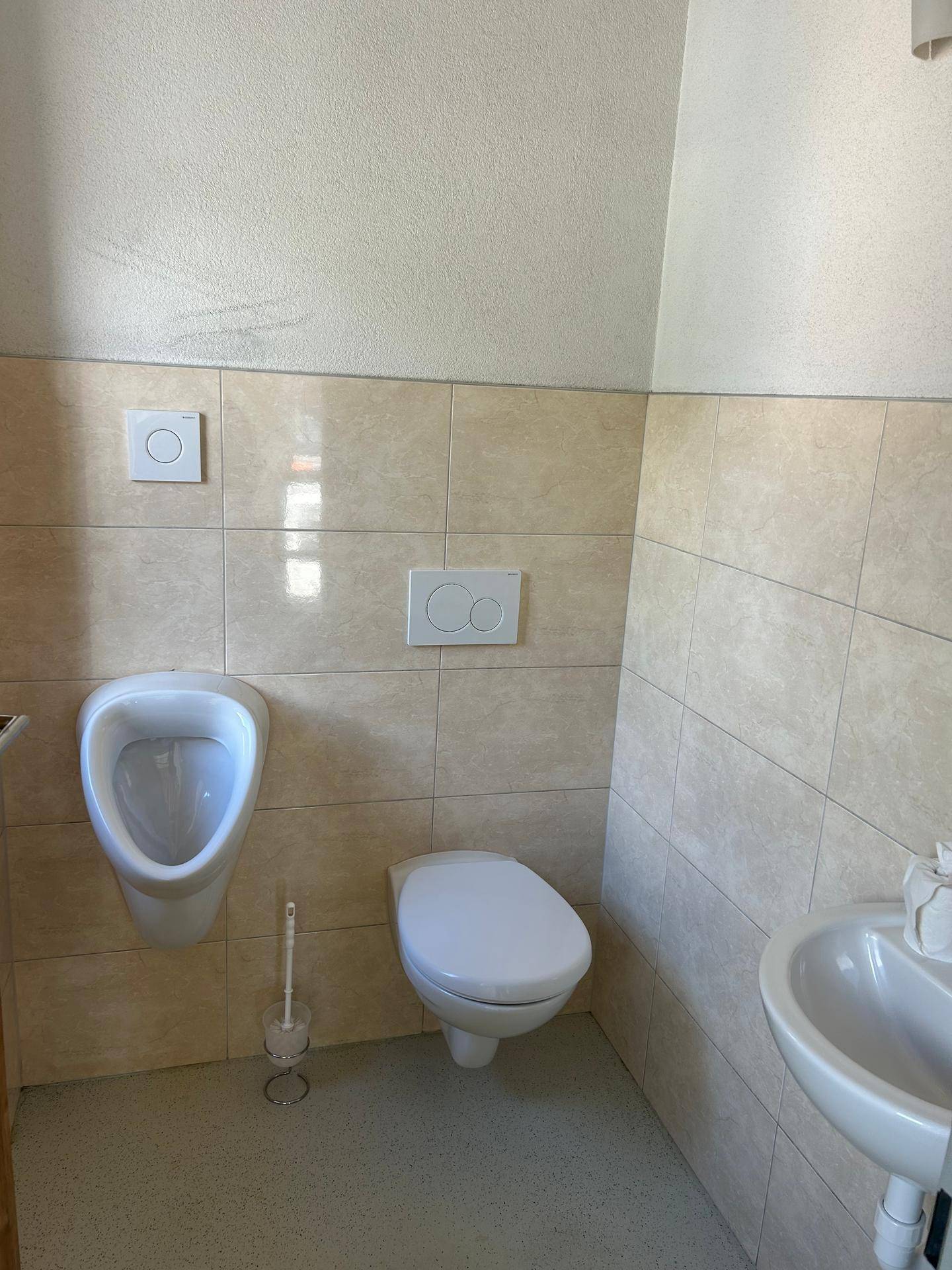 Gefliestes Badezimmer mit Urinal, Toilette und Waschbecken in hellen Farbtönen.