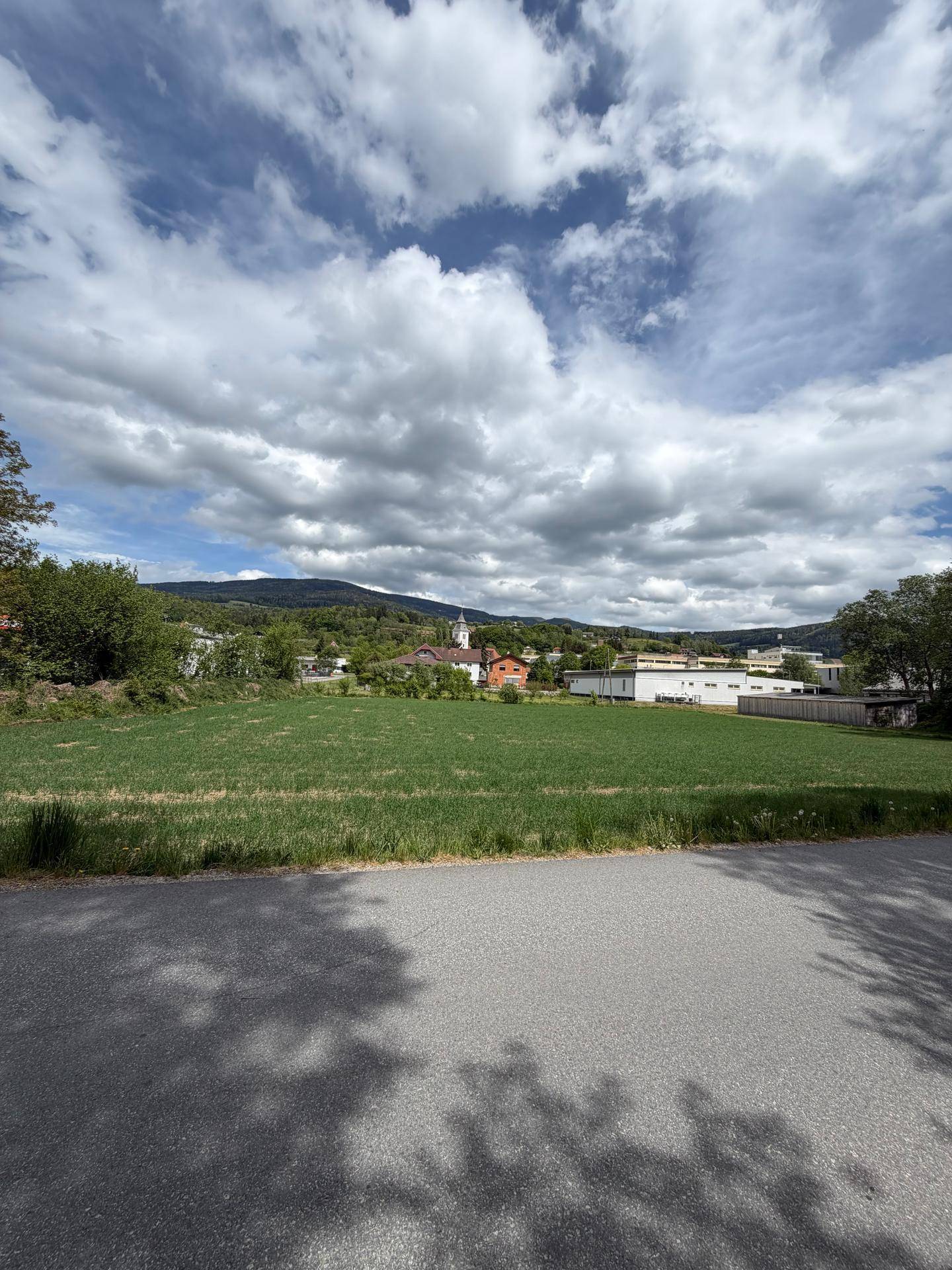 Blick über ein grünes Feld auf ein Dorf mit Kirche und Berge, im Vordergrund eine asphaltierte Straße.