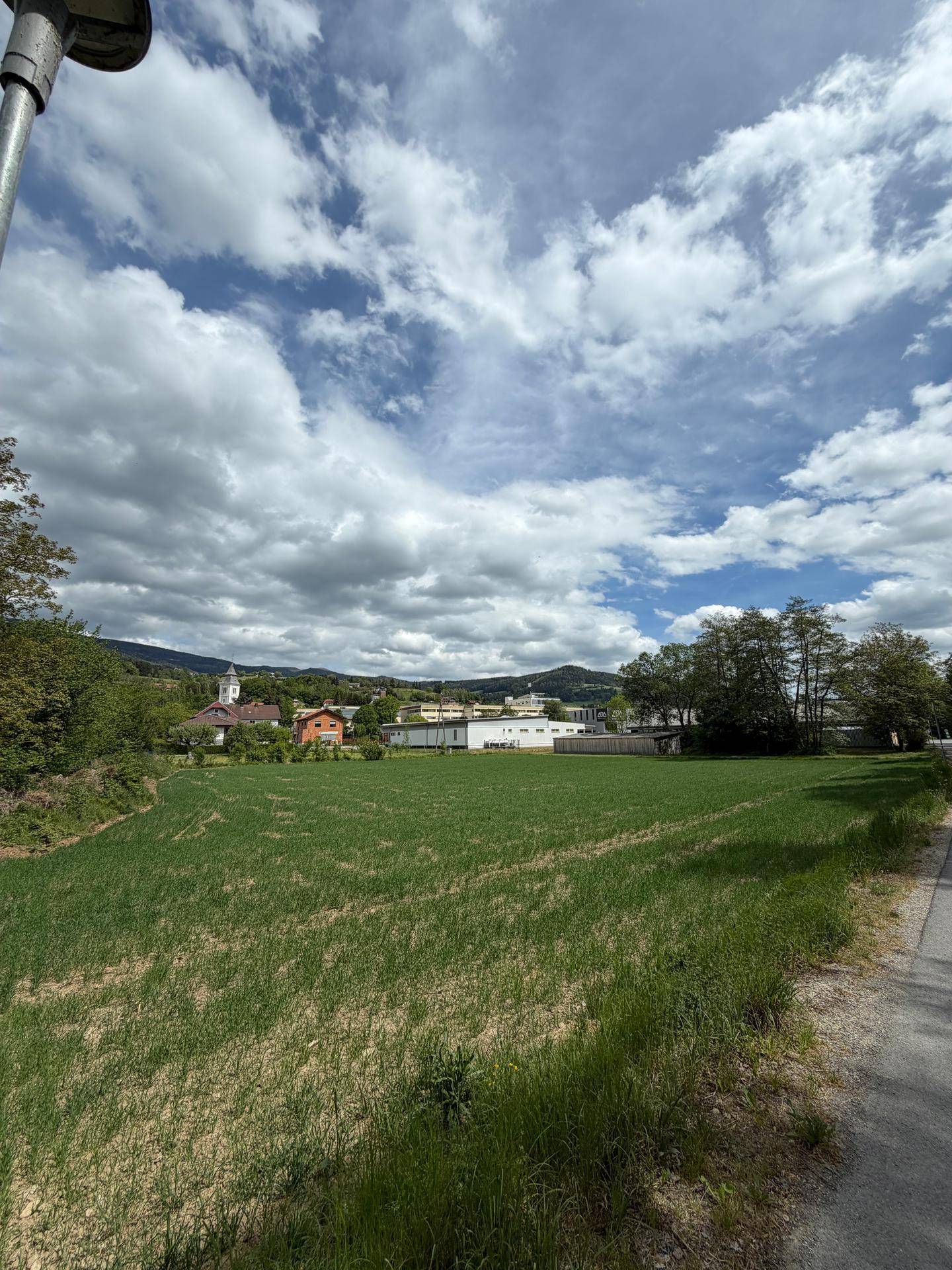 Panoramablick über ein weites Feld auf eine ländliche Siedlung und bewaldete Hügel unter blauem Himmel.