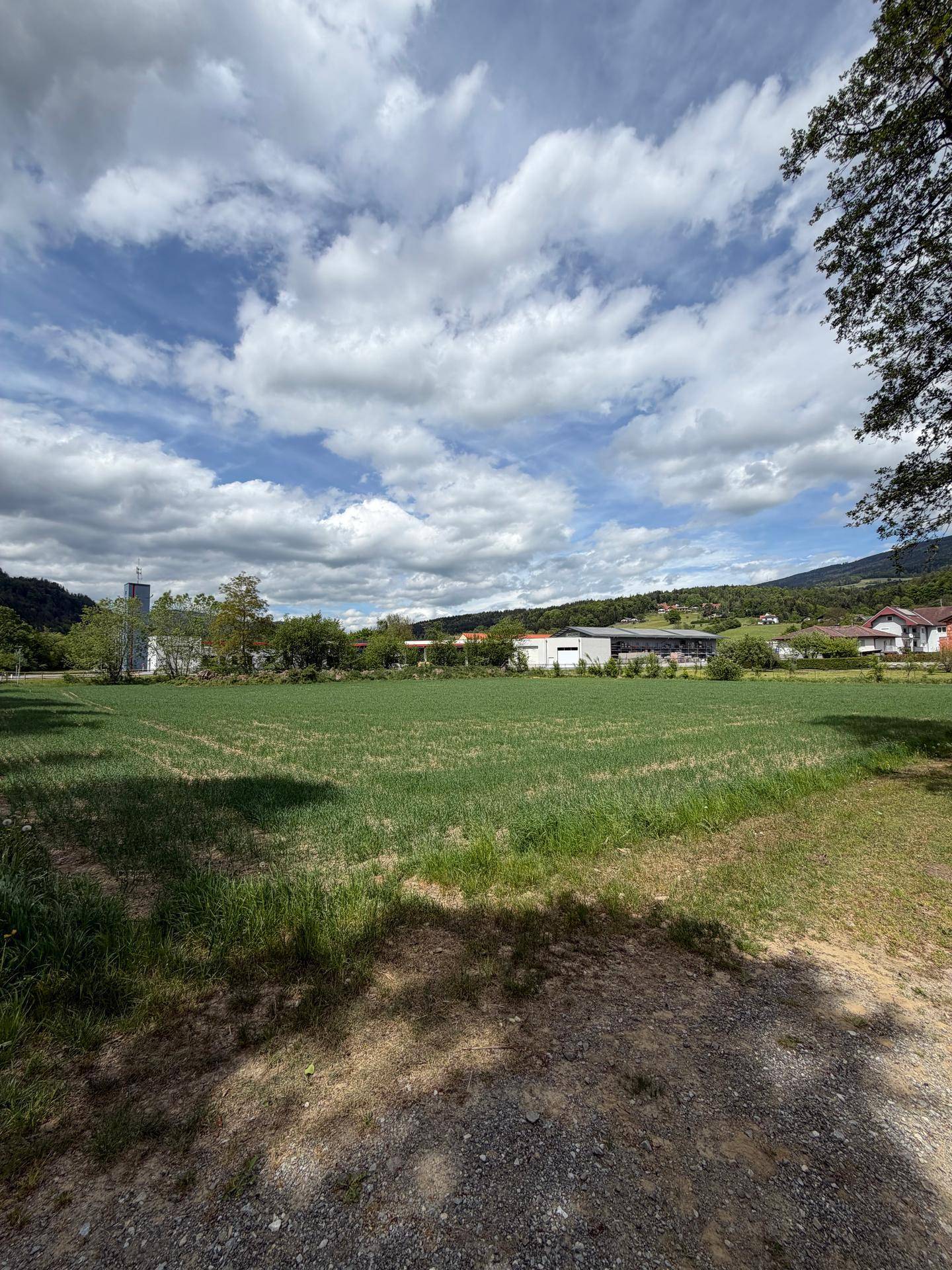 Weitläufiges grünes Feld mit Blick auf eine kleine Ortschaft und die umliegende Berglandschaft.