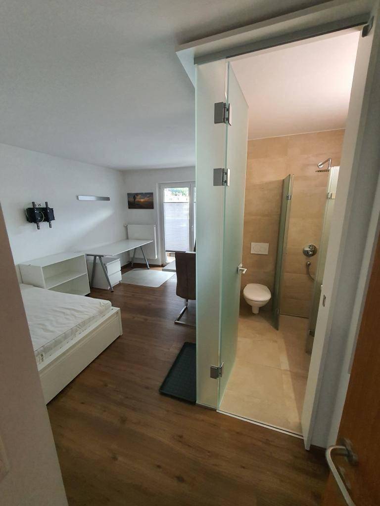 Helles Studioapartment mit Schlafbereich, Schreibtisch und angrenzendem Badezimmer hinter einer Glastür.