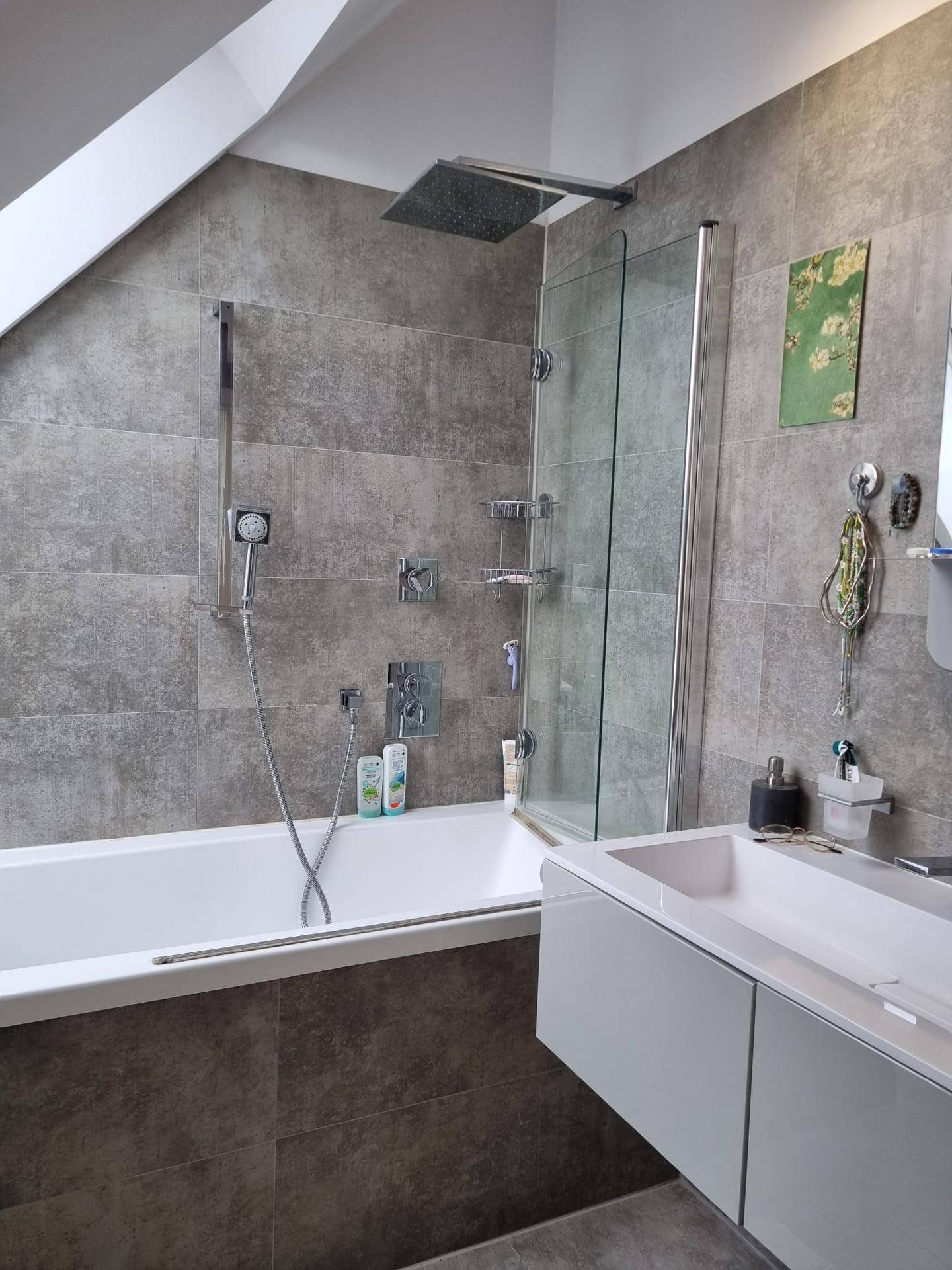 Badezimmer mit Badewanne, integrierter Dusche und modernem Waschtisch, gefliest in Grautönen.