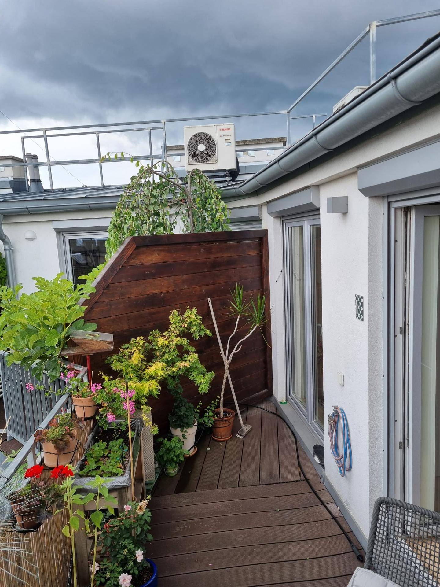 Begrünte Dachterrasse mit Holzbelag, Pflanzen und einer Klimaanlage an der Fassade.