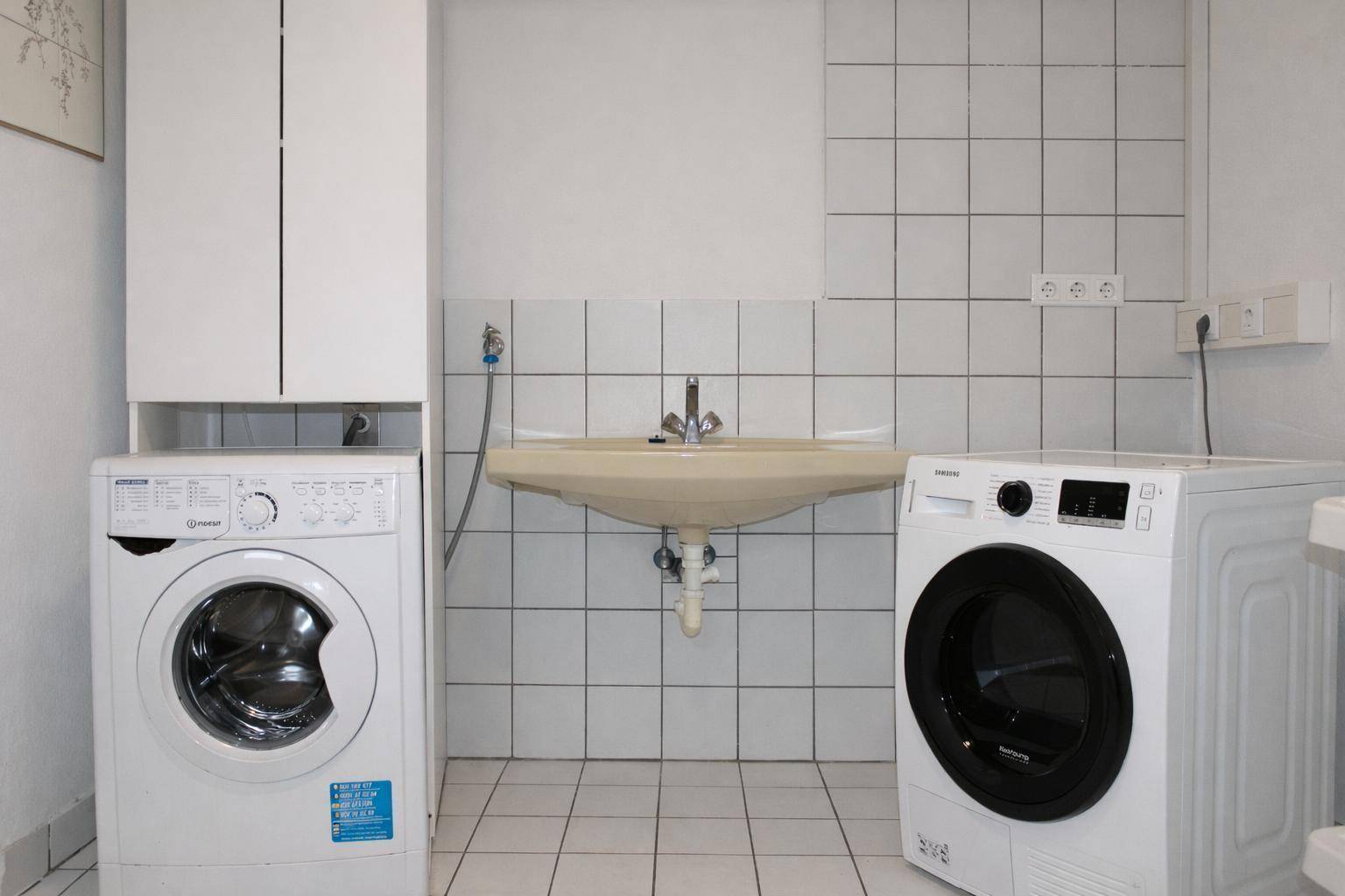 Waschküche mit Waschmaschine, Trockner, Waschbecken und weißen Wandfliesen.