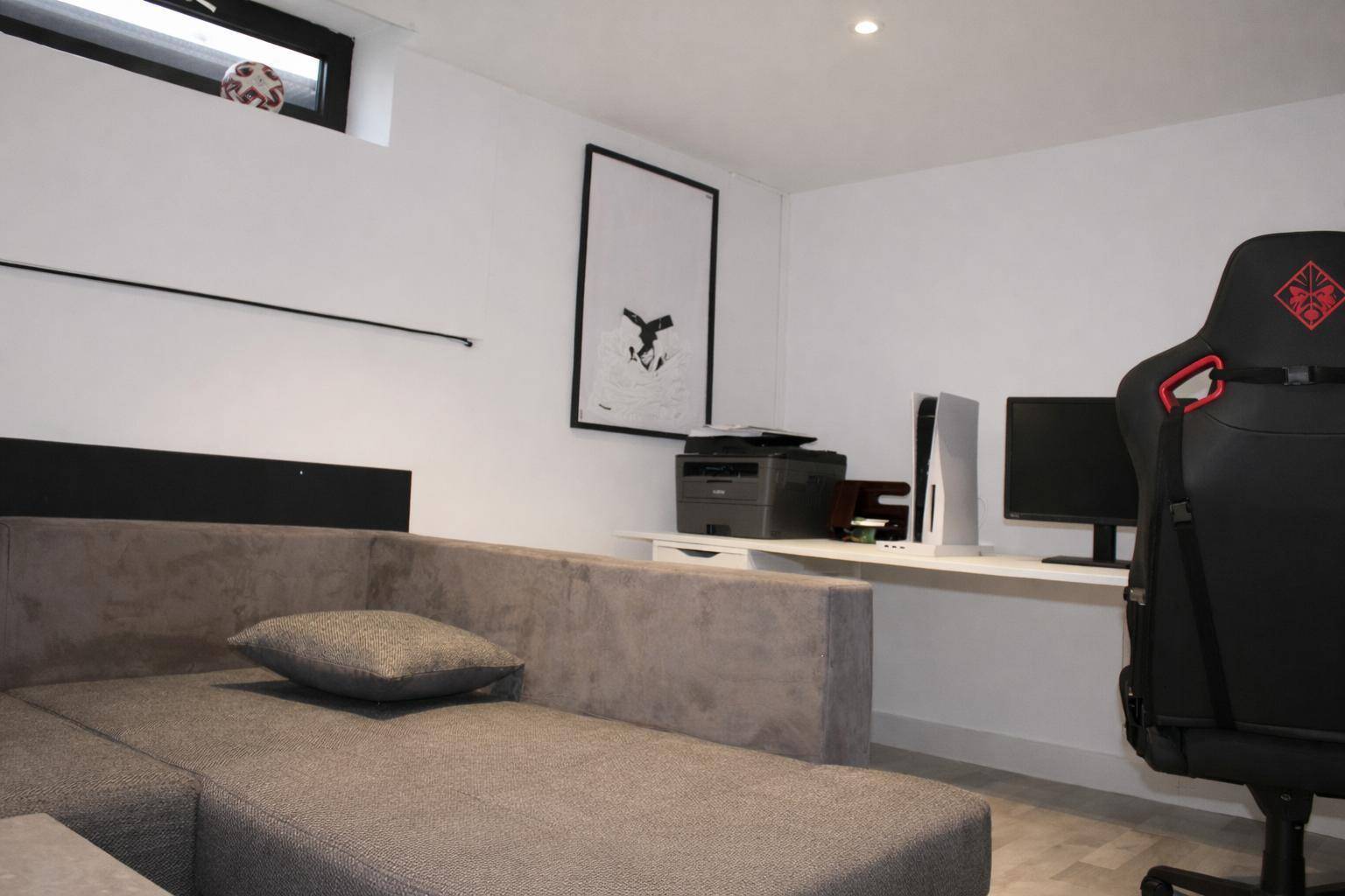 Hobbyraum im Keller mit Sofa, Schreibtisch, Gaming-Stuhl und kleinem Fenster.