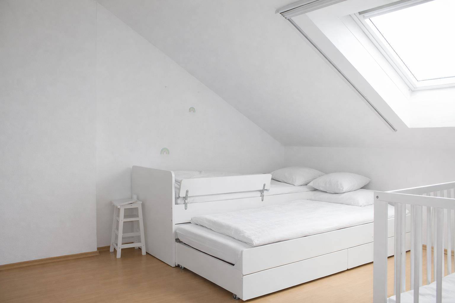 Kinderzimmer im Dachgeschoss mit weißem Bett, Babybett und großem Dachfenster.