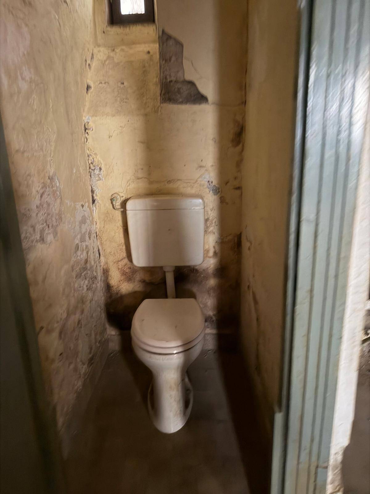 Kleine Toilette in schlechtem Zustand mit sichtbaren Abnutzungsspuren und einem kleinen Fenster.