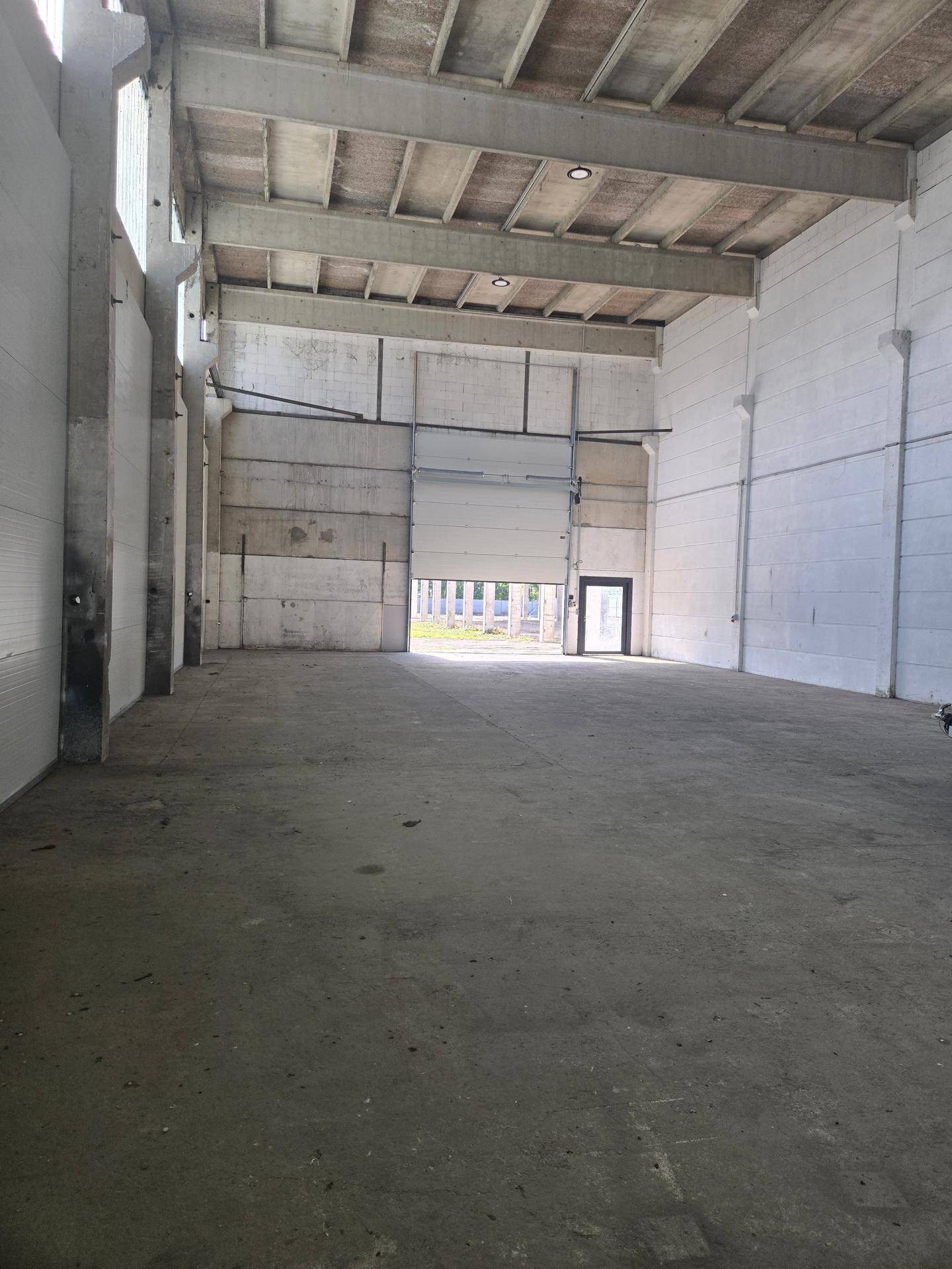 Weitläufige Industriehalle mit hoher Decke, Betonkonstruktion und großem Zugangstor mit Nebentür am Ende des Raumes.
