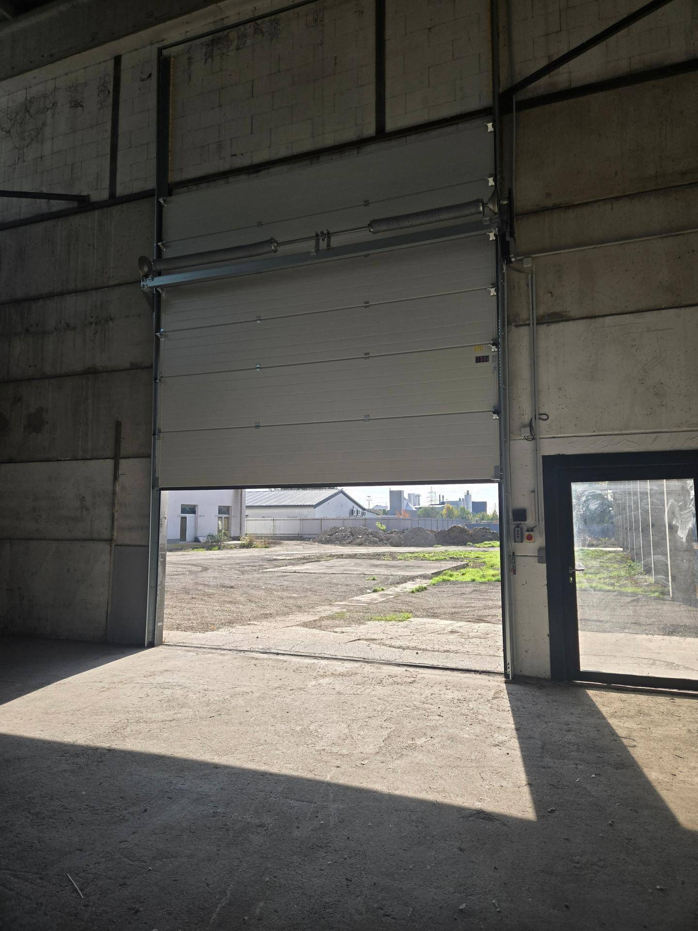 Blick aus der Halle durch das teilweise geöffnete Rolltor auf den Außenbereich und eine separate Tür daneben.