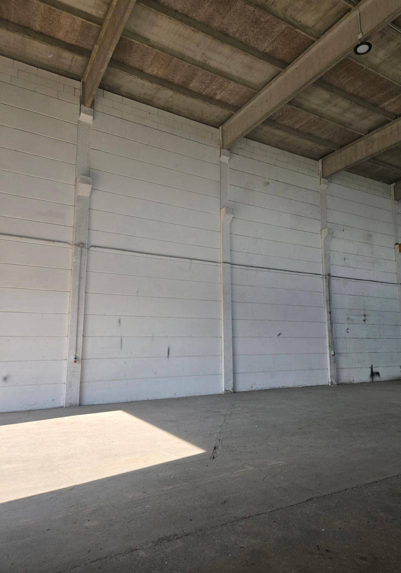 Leere Lagerhalle mit Betonwänden und -boden, sowie sichtbaren Deckenbalken in einem funktionalen Industriestil.
