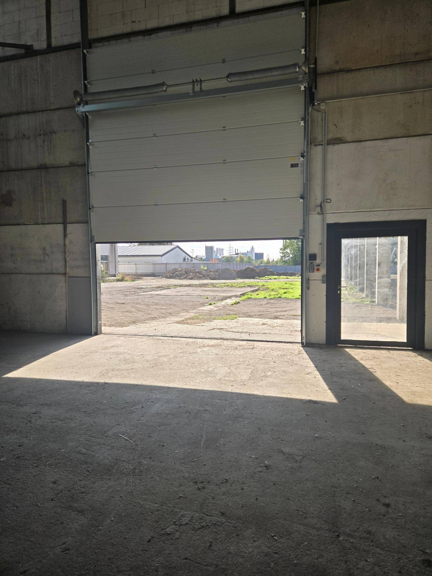 Innenansicht der Halle mit Blick durch das geöffnete Rolltor auf den Hof und eine Nebentür für den Zugang.