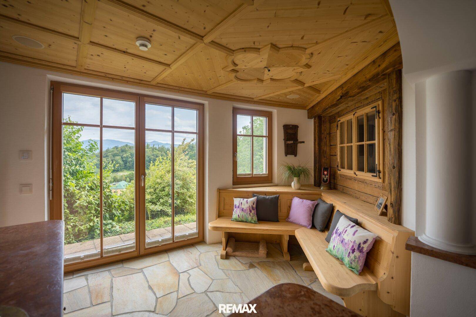 Rustikaler Wohnbereich mit Holzbalkendecke, Steinboden und großen Fenstern mit Blick ins Grüne.