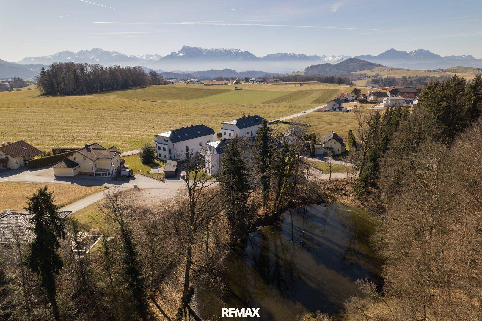 Luftaufnahme einer ländlichen Gegend mit Häusern, Feldern, einem Teich und Bergen im Hintergrund.