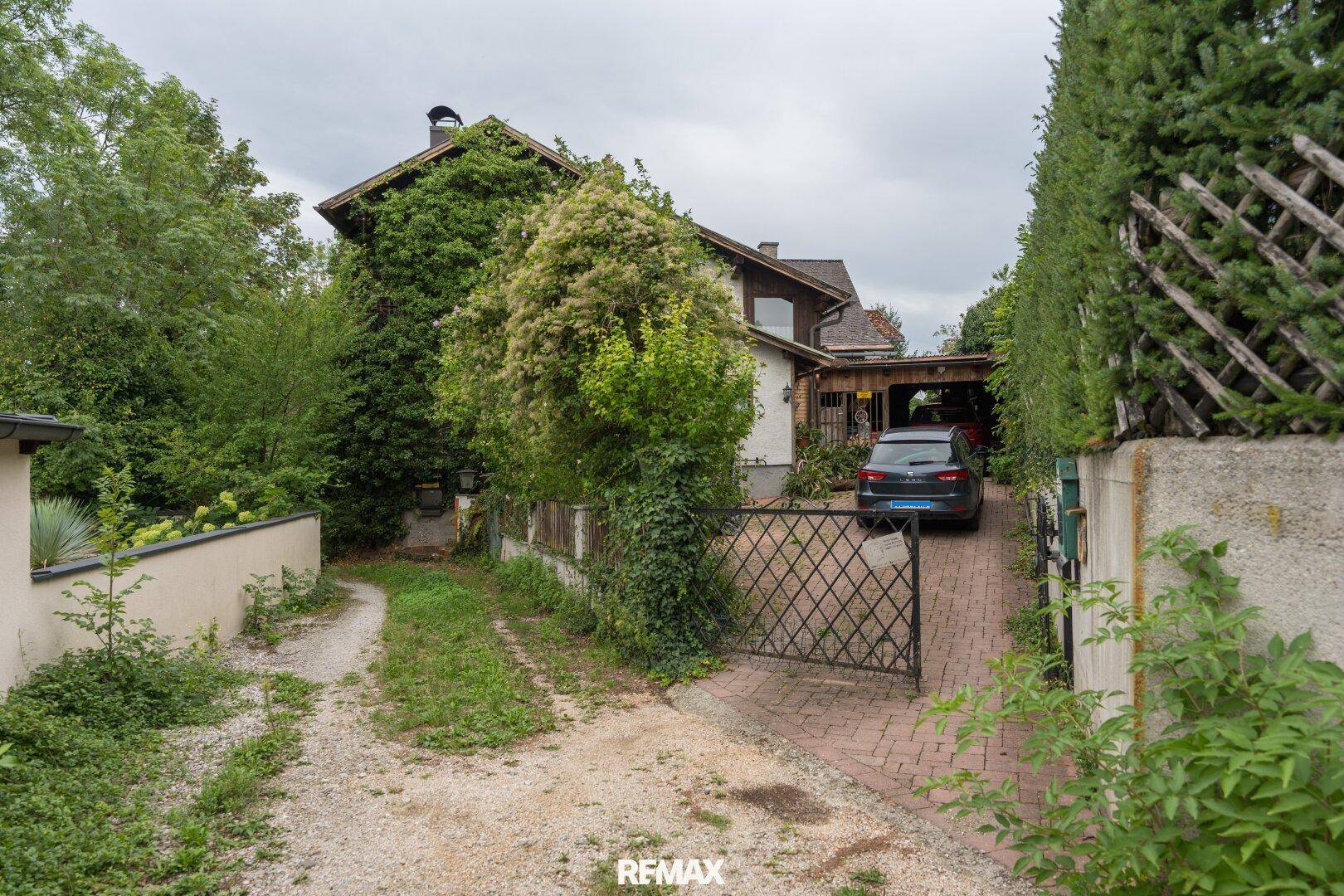 Einfahrt zum Haus mit Carport, umgeben von dichtem Grün und einem schmiedeeisernen Tor.