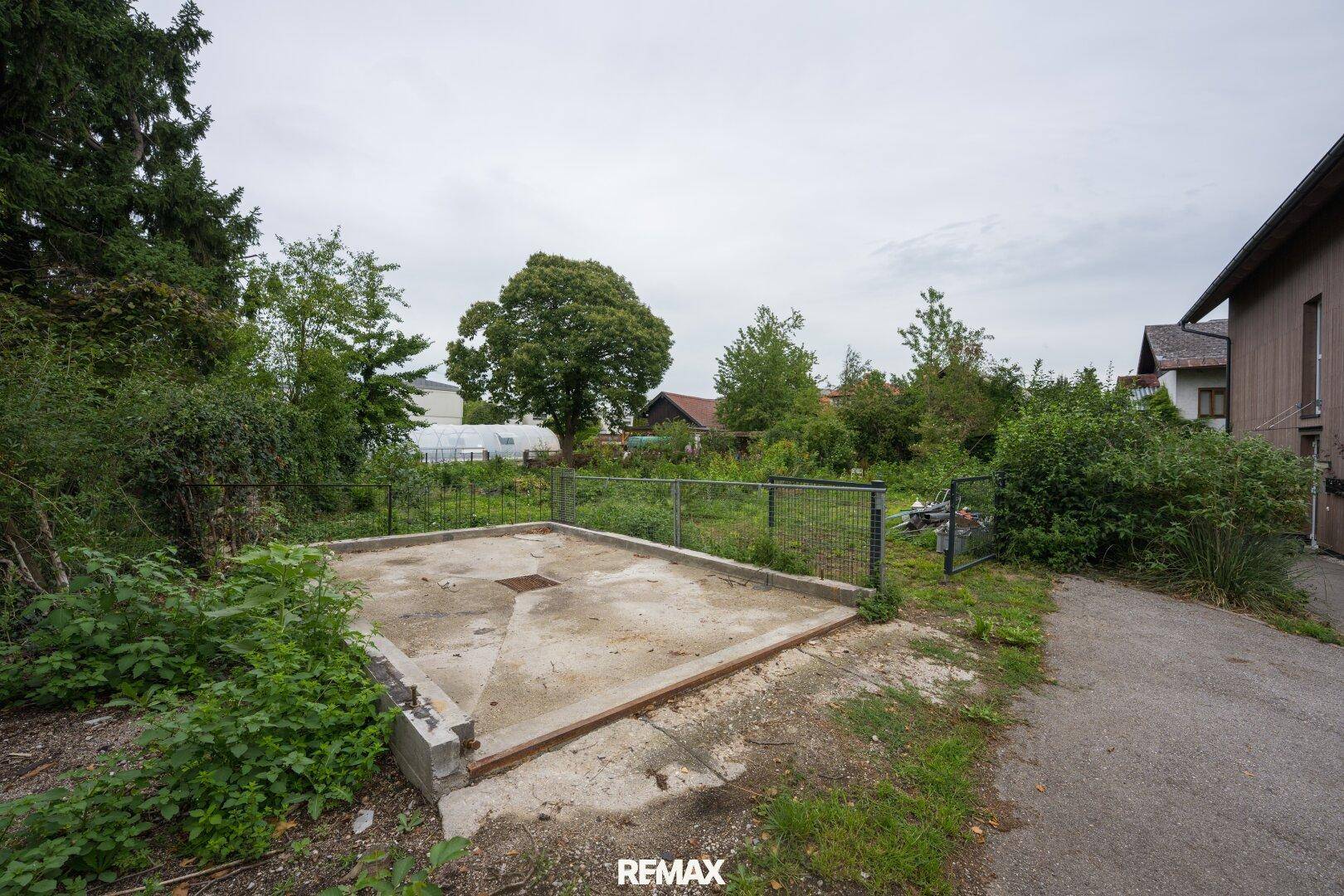 Betonfundament auf dem Grundstück, umgeben von üppiger Vegetation und einem Zaun.