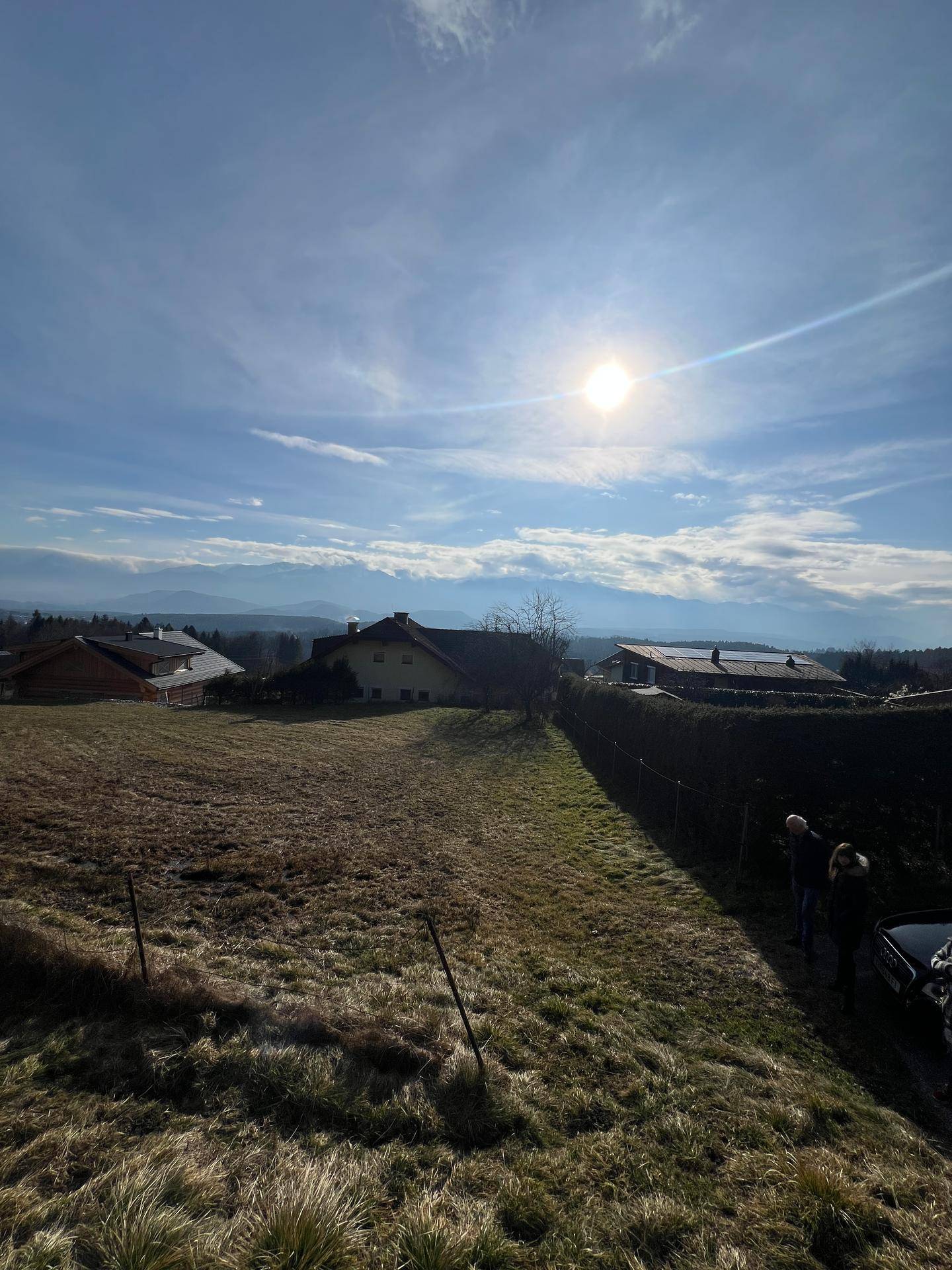 Weitläufiges Grundstück mit trockener Wiese und Häusern im Hintergrund, sonniger Wintertag mit Bergblick.