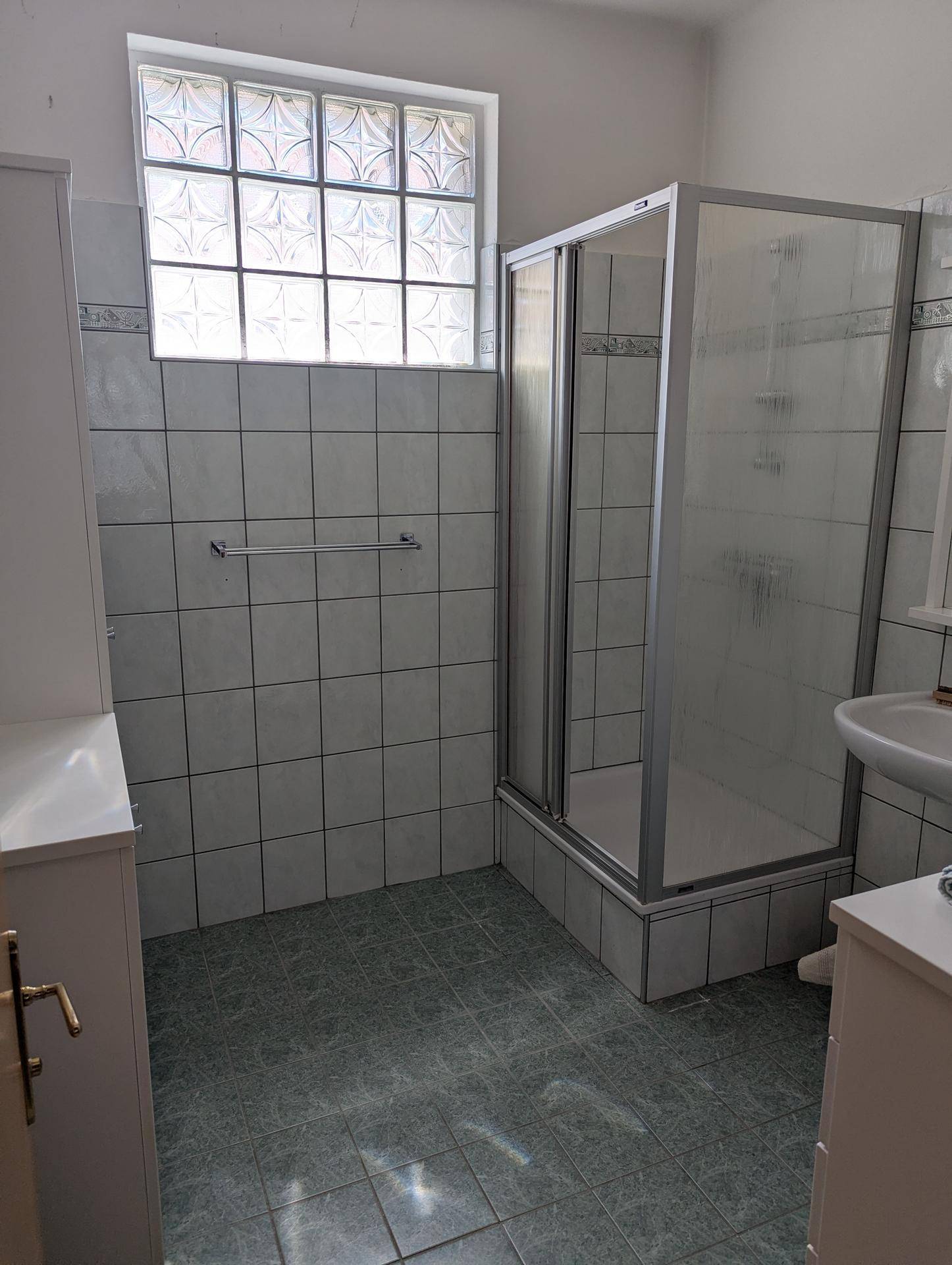 Badezimmer mit Duschkabine, weißen Fliesen und Fenster aus Glasbausteinen.