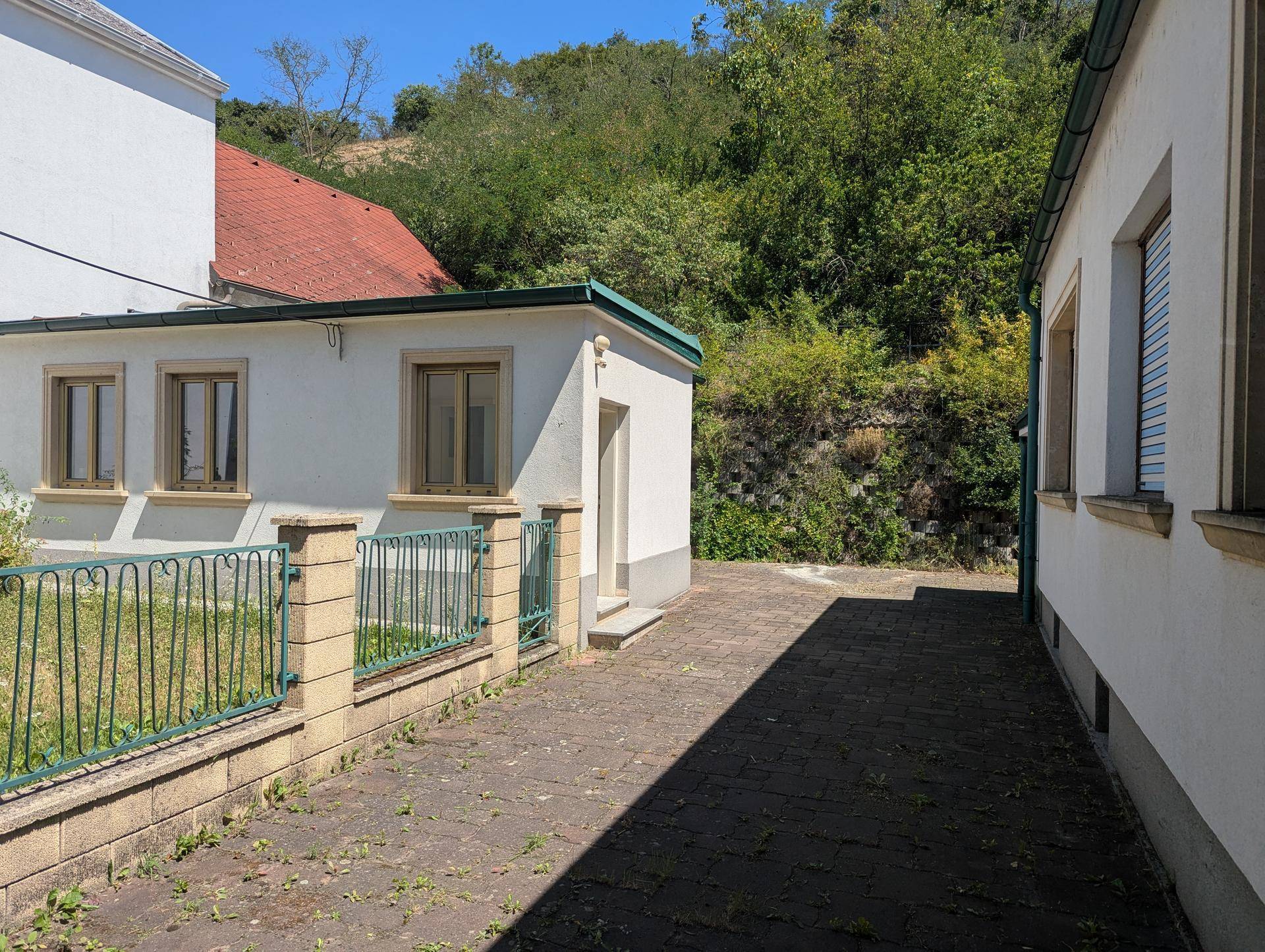 Seitenansicht eines Gebäudes mit gepflastertem Weg und Blick auf bewaldeten Hang.