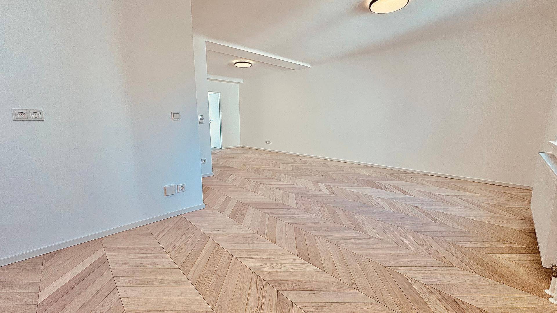 Weitläufiger, heller Wohnbereich mit elegantem Fischgrätparkett und schlichten Wänden.