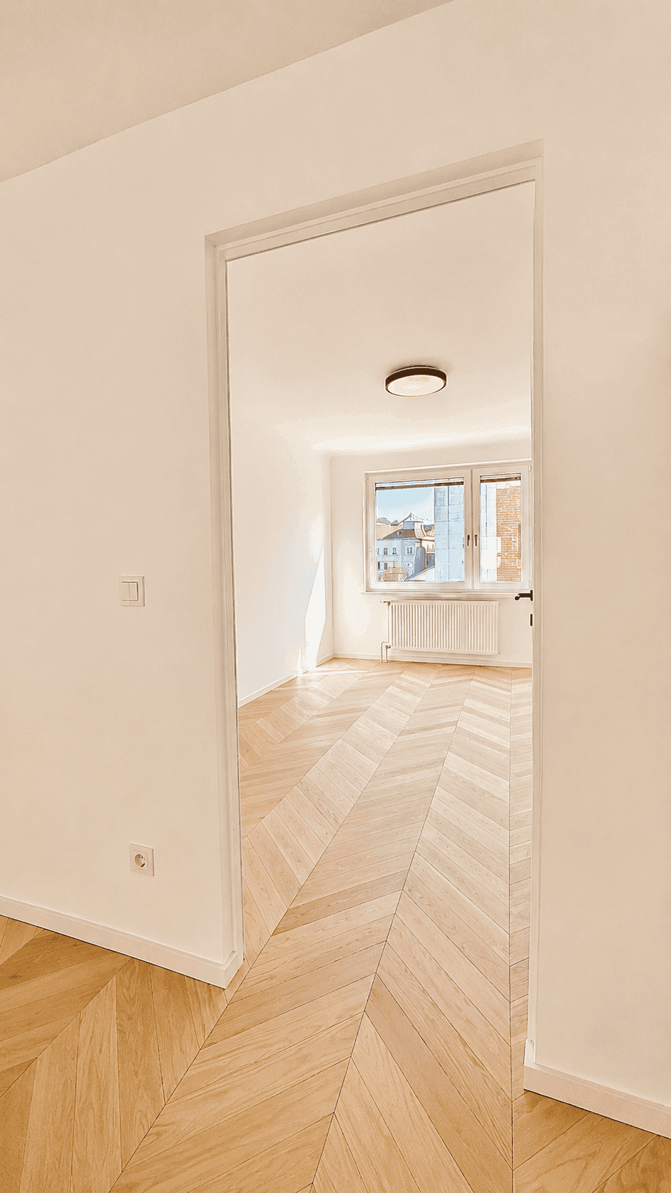 Blick durch eine Tür in ein helles Zimmer mit Fischgrätparkett und großen Fenstern.