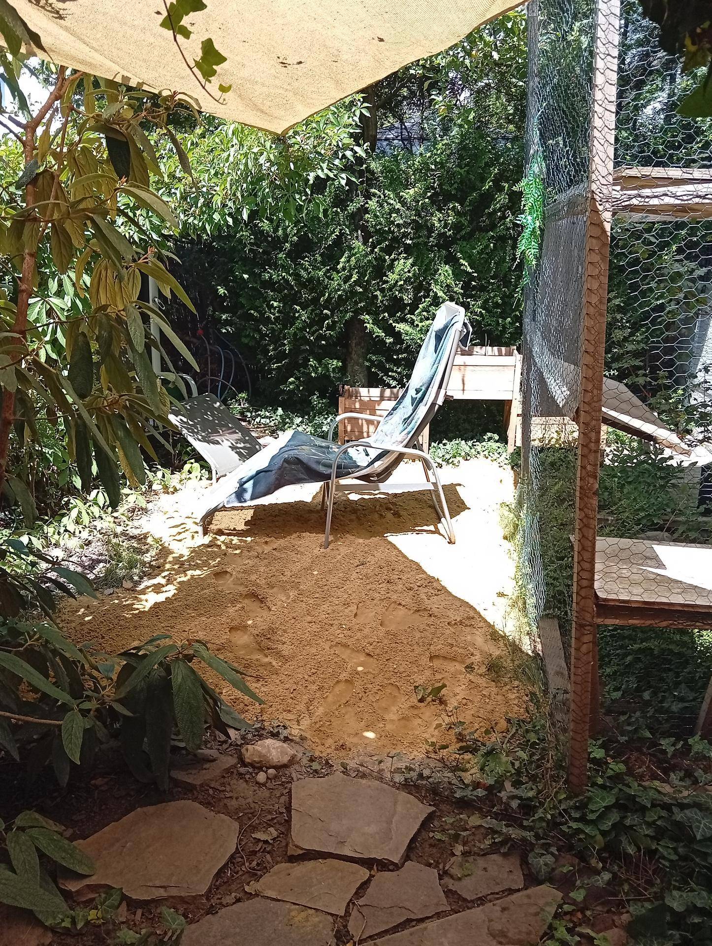 Entspannter Gartenbereich mit Liegestuhl, Sandfläche und üppiger Vegetation unter einem Sonnensegel.