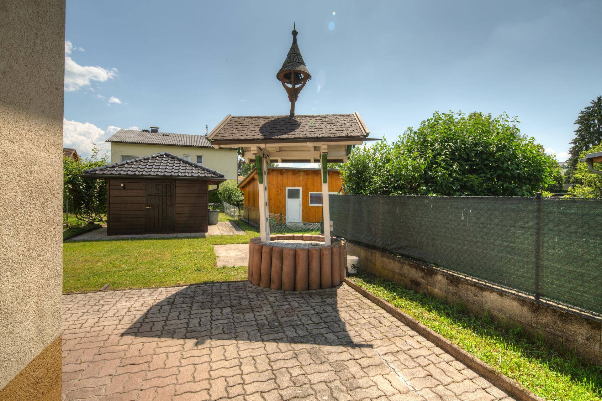 Dekorativer Brunnen mit Holzdach und gepflastertem Bereich im sonnigen Garten.