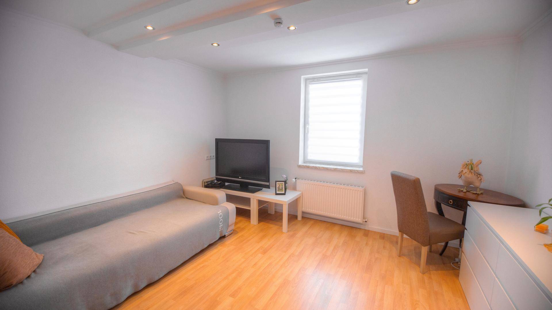 Helles Wohnzimmer mit Sofa, Fernseher und Laminatboden, gut beleuchtet durch ein Fenster.
