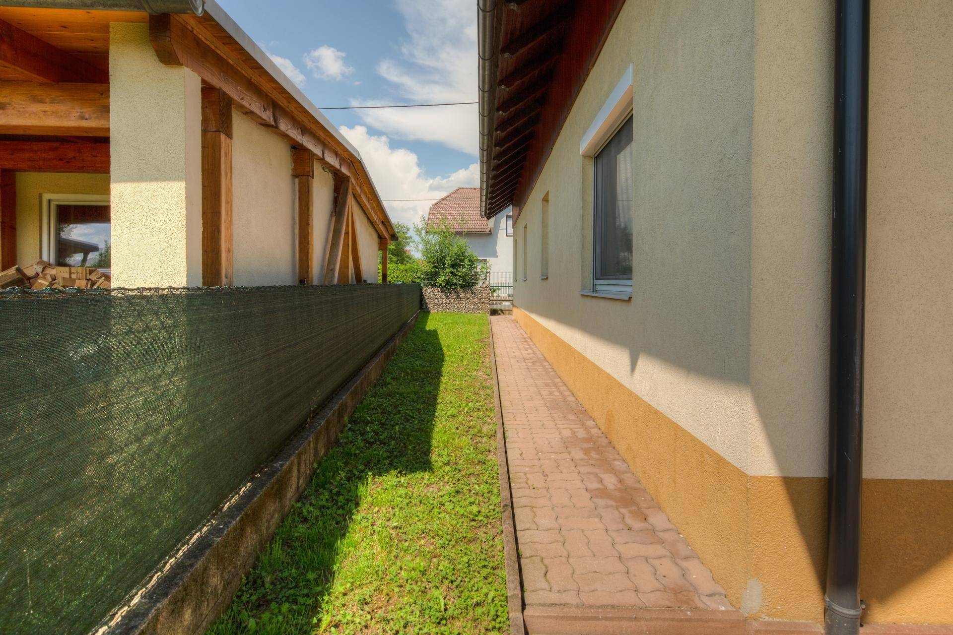 Schmaler, gepflasterter Weg entlang der Hauswand mit Rasenfläche und Zaun.