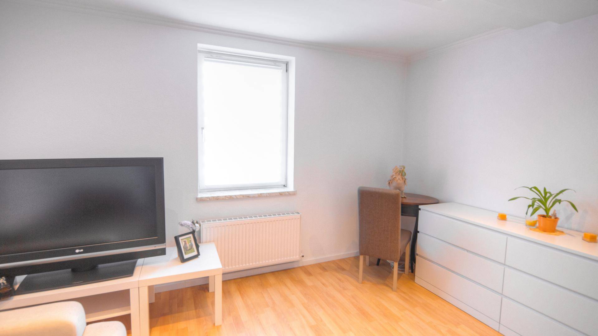 Wohnzimmer mit Fernseher, Kommode und Fenster, das für ausreichend Tageslicht sorgt.