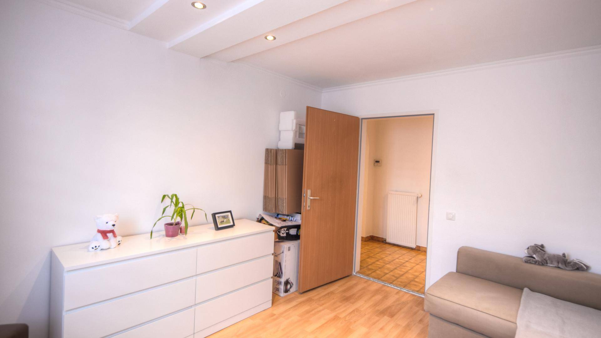 Wohnzimmer mit weißer Kommode, Holzboden und offenem Zugang zu einem weiteren Raum.