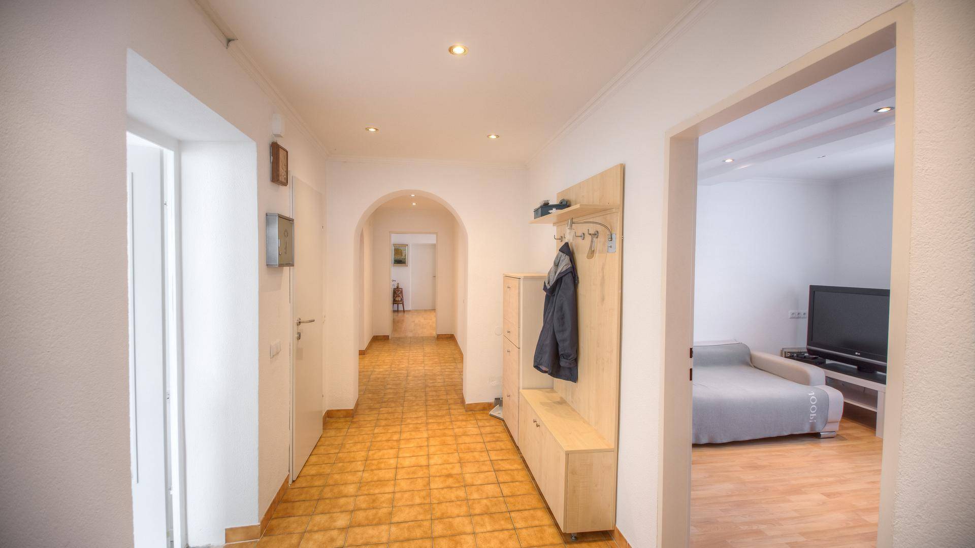 Langer Flur mit hellen Fliesen, Holzboden im angrenzenden Raum und Garderobe.