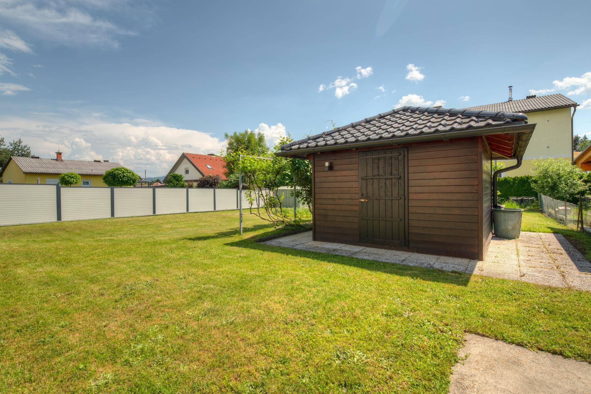 Holzgartenhaus mit gepflastertem Zugangsweg und umgebender Rasenfläche im Sonnenschein.