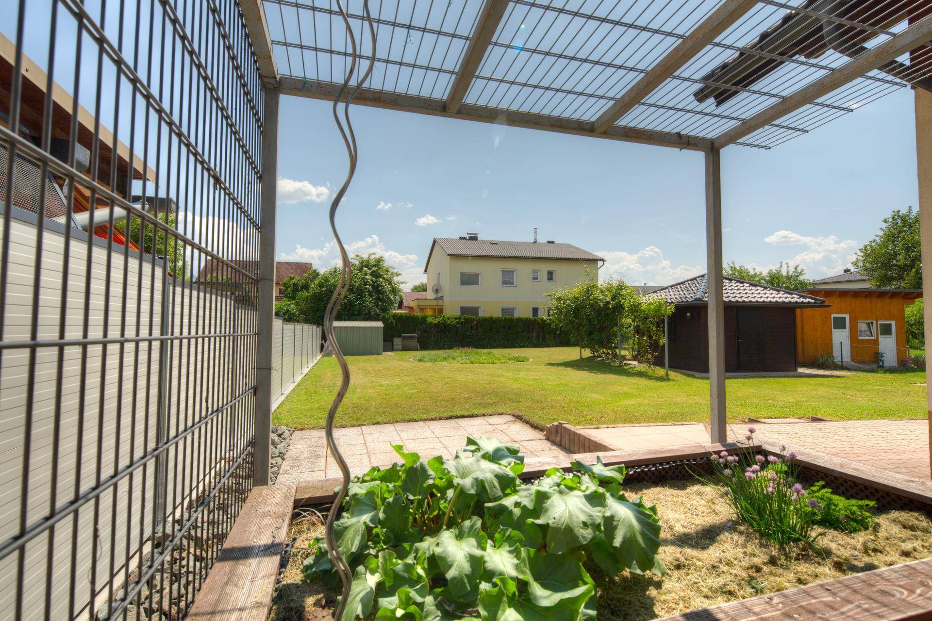 Weitläufiger Garten mit gepflegtem Rasen, Hochbeet und einer Metallpergola unter blauem Himmel.