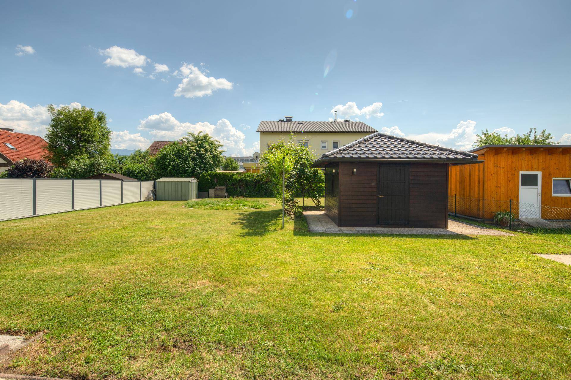 Weitläufiger Garten mit Rasenfläche, zwei Gartenhäusern und einem Zaun im Hintergrund.