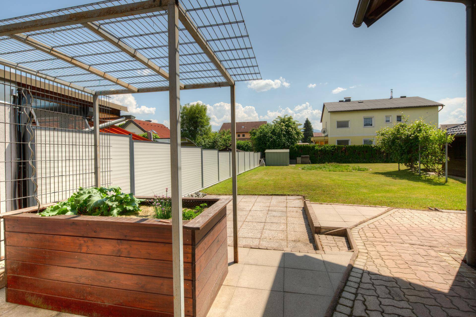 Blick in den Garten mit Hochbeet, Rasenfläche und einer Metallpergola im Sonnenschein.