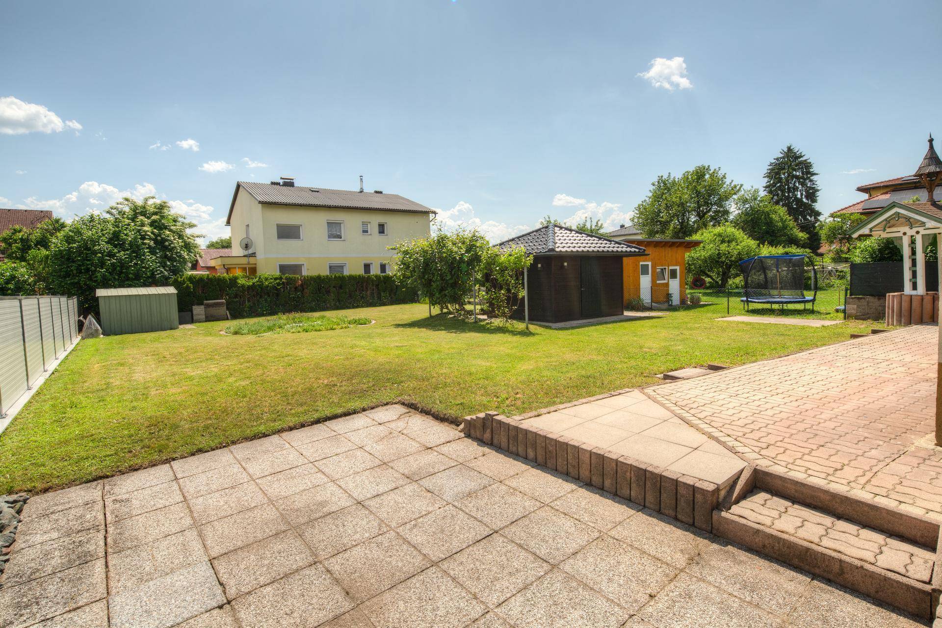 Große Rasenfläche im Garten mit gepflasterter Terrasse und Blick auf Nachbarhäuser.