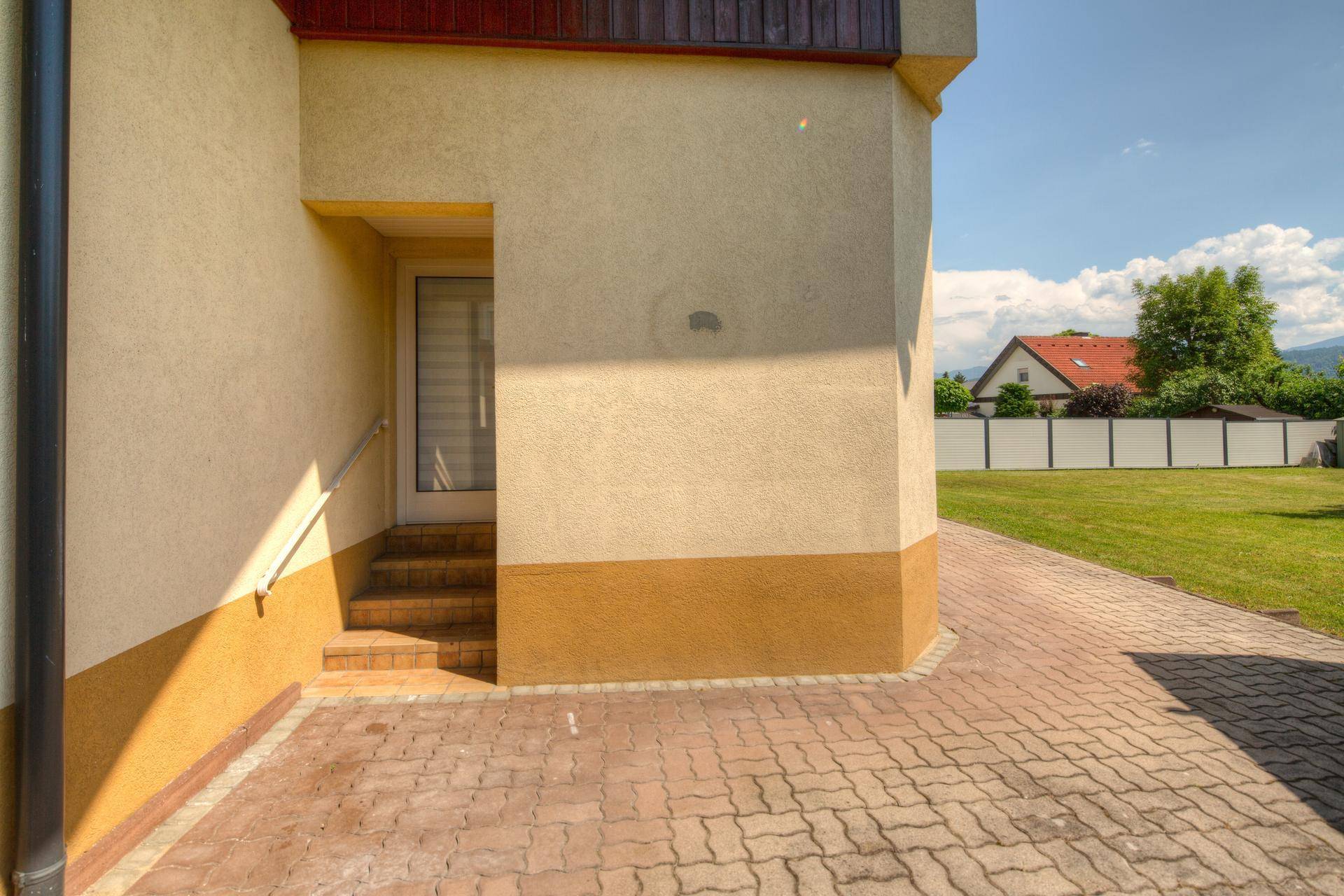 Eingangsbereich des Hauses mit Treppe und gepflastertem Vorplatz an einem sonnigen Tag.