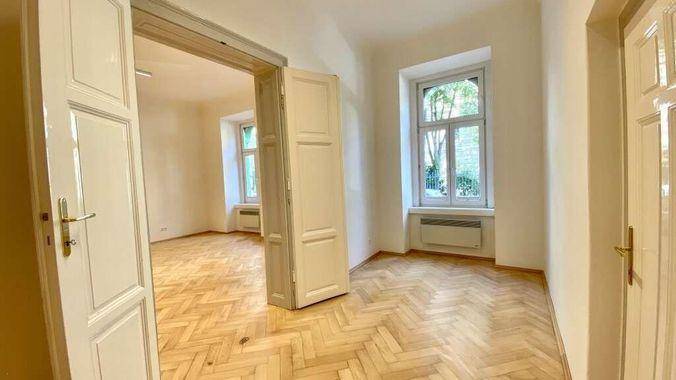 Geräumiger Raum mit klassischem Fischgrätparkett, hohen Decken und einem Fenster mit Blick ins Grüne.