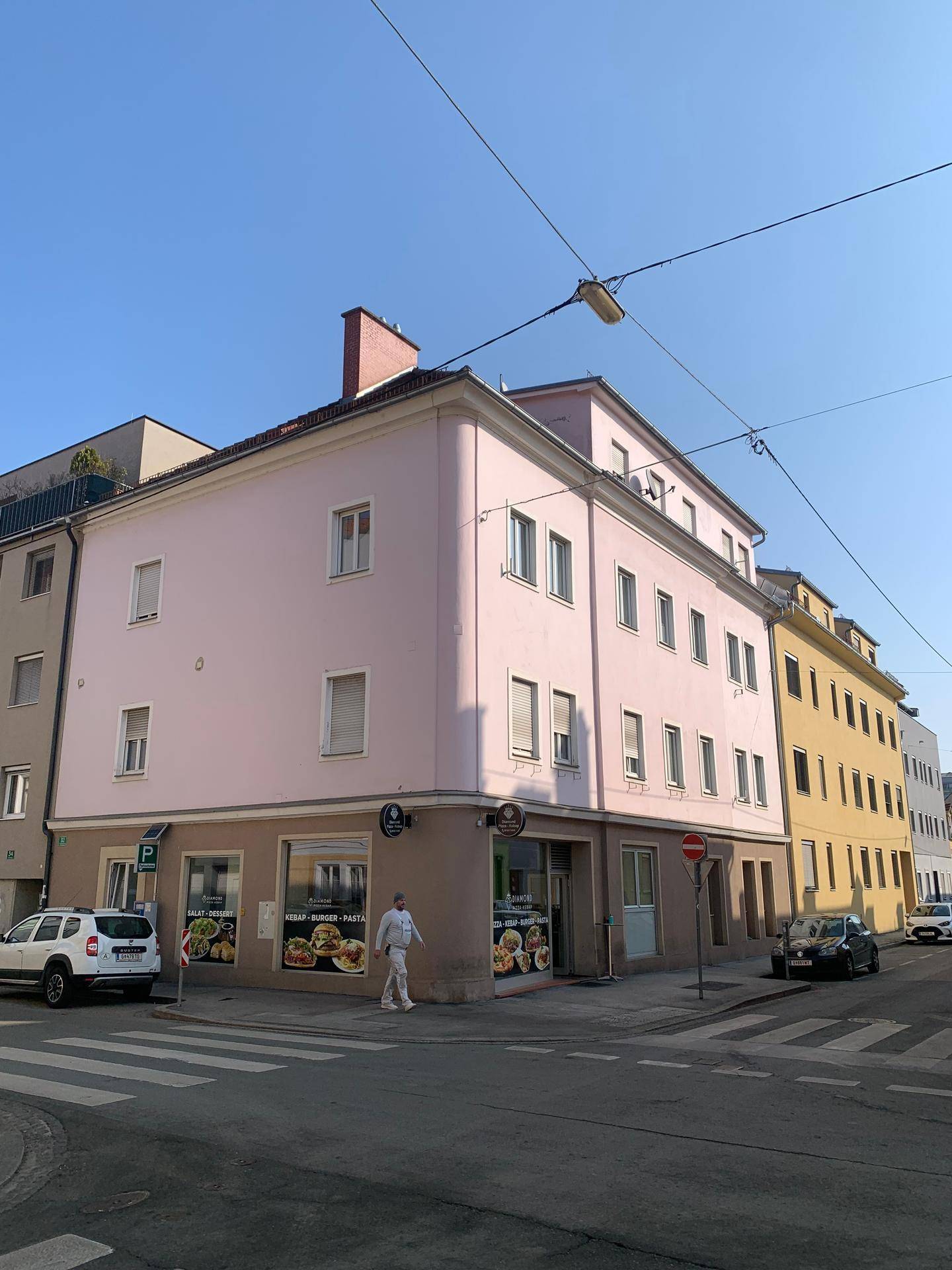 Eckgebäude mit rosa Fassade und Geschäften im Erdgeschoss an einer städtischen Kreuzung.