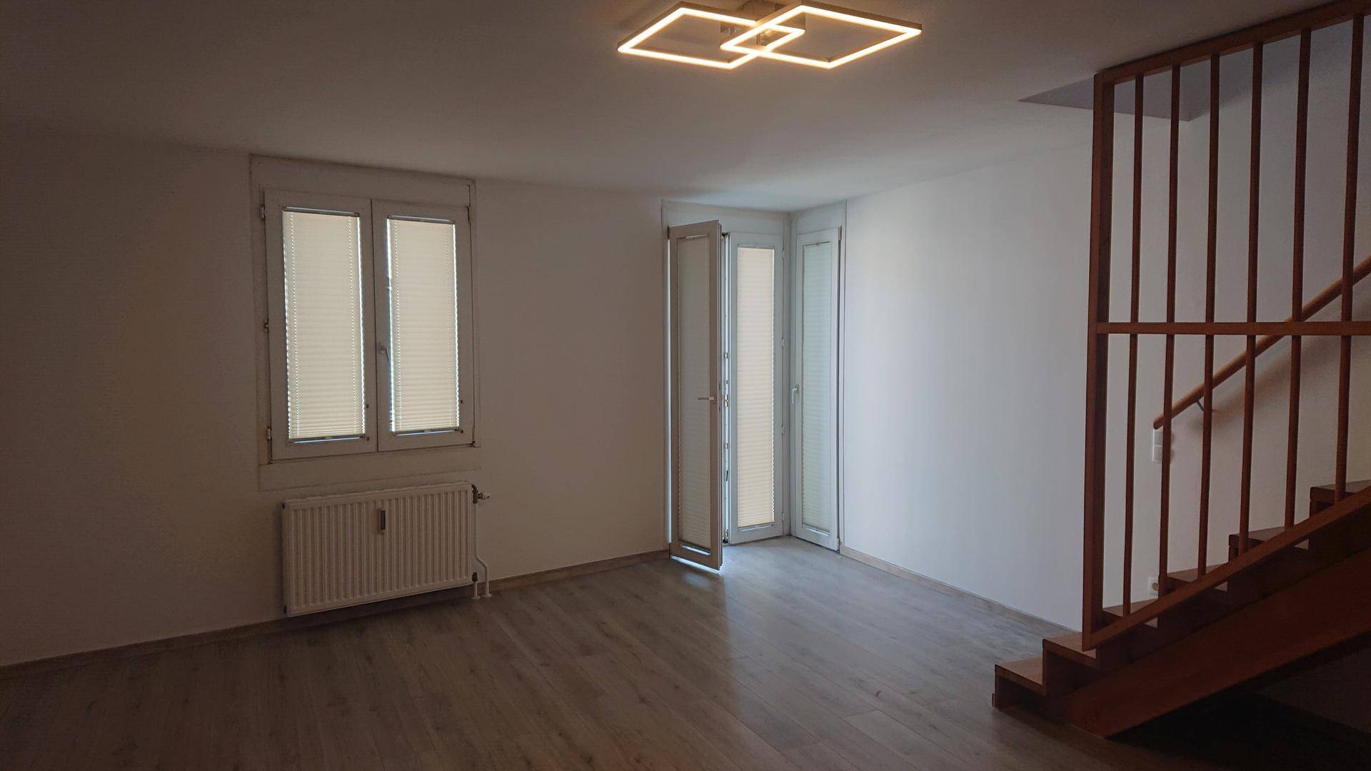 Heller Wohnraum mit modernem Deckenlicht, Laminatboden und Zugang zur Holztreppe.